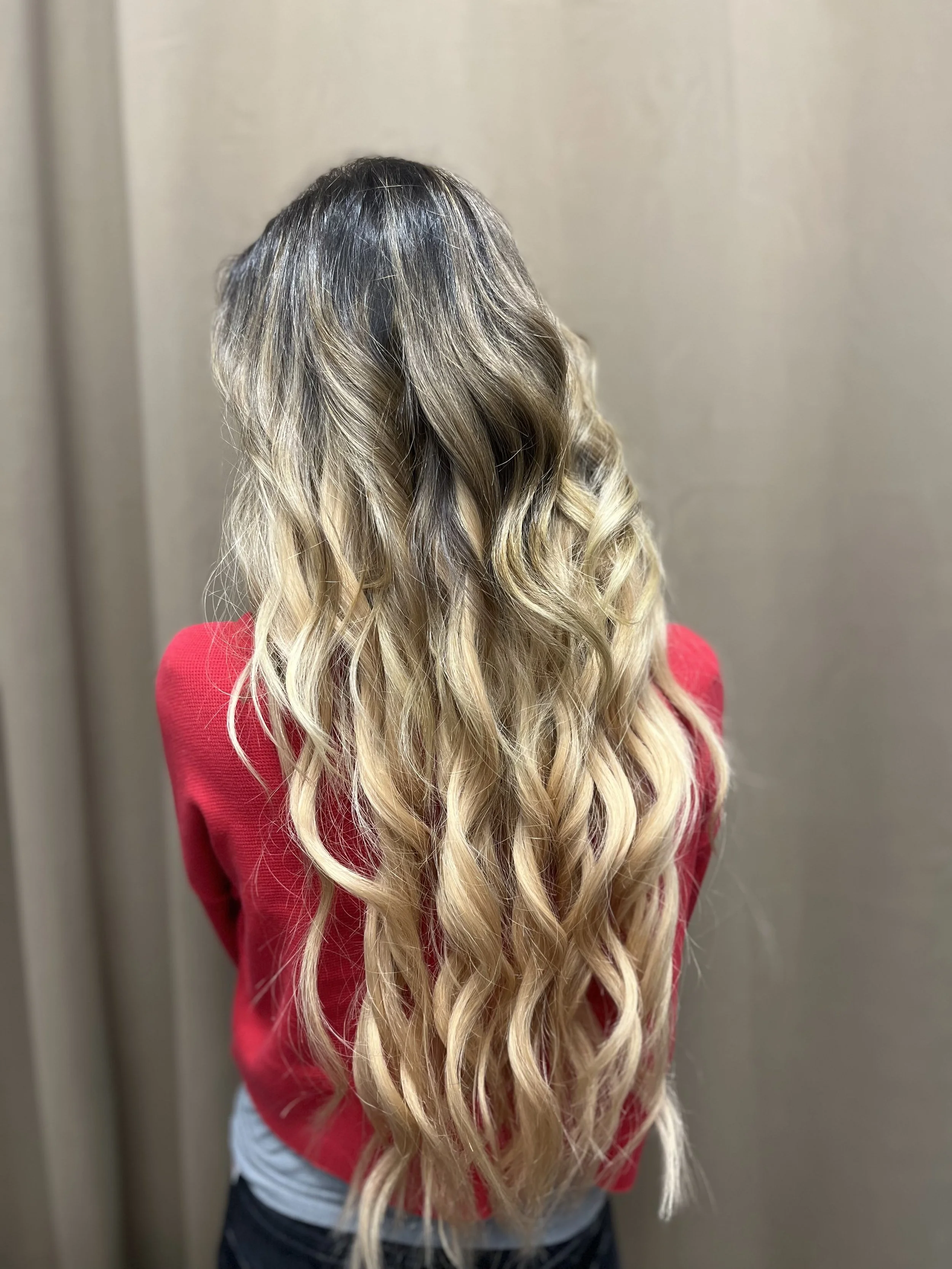 Back of a woman with long, wavy blonde hair, wearing a red top, standing against a beige wall.