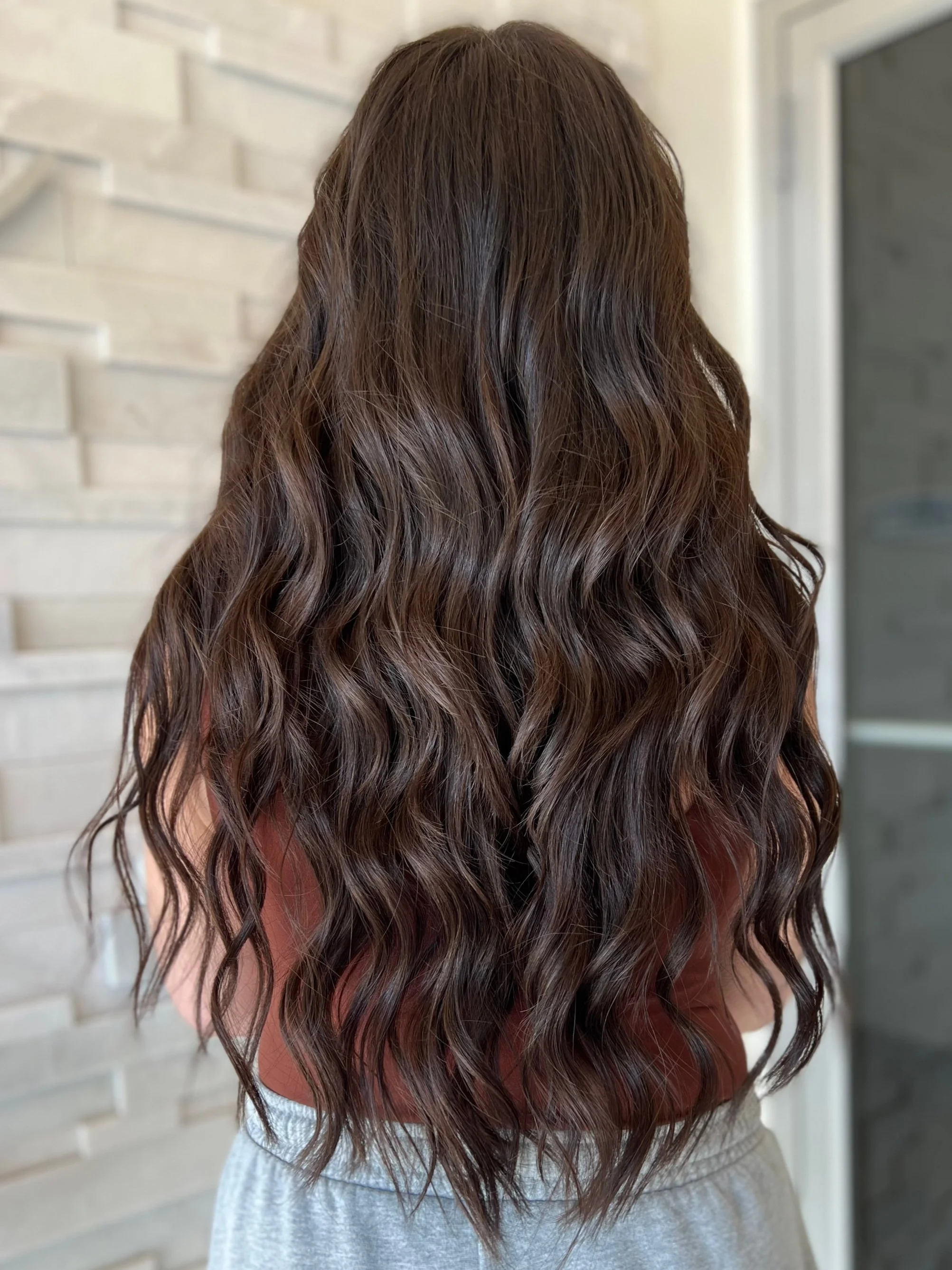 Woman with long, wavy brown hair standing indoors, facing a textured light-colored wall.