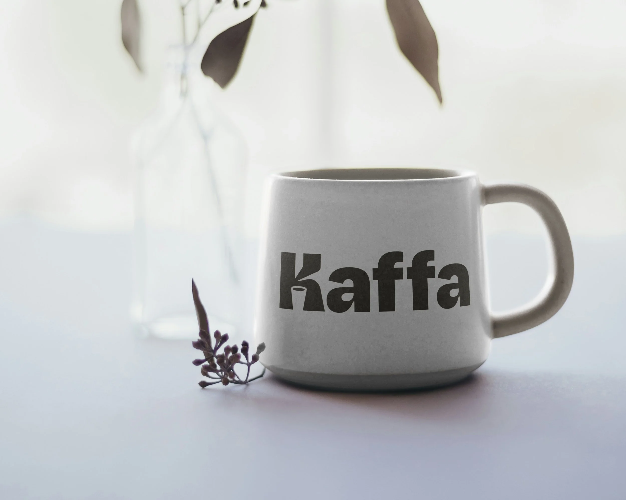 A white coffee mug with the word 'Kaffa' in bold black letters, placed on a white surface with a small sprig of purple flowers beside it. A glass vase with dark leaves is in the background.
