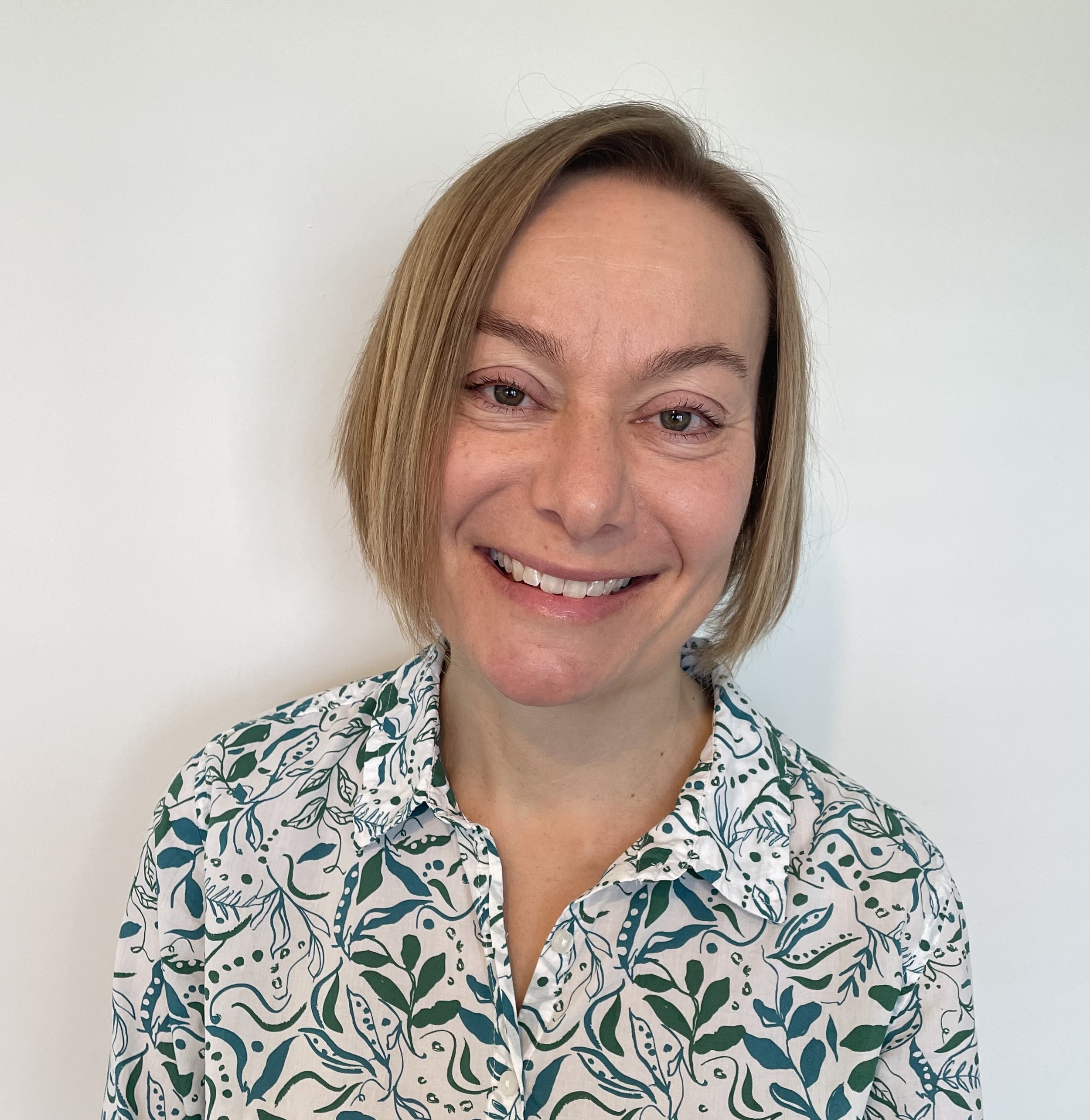 A woman with short blonde hair smiling, wearing a white shirt with green and navy floral pattern, standing against a plain white wall.