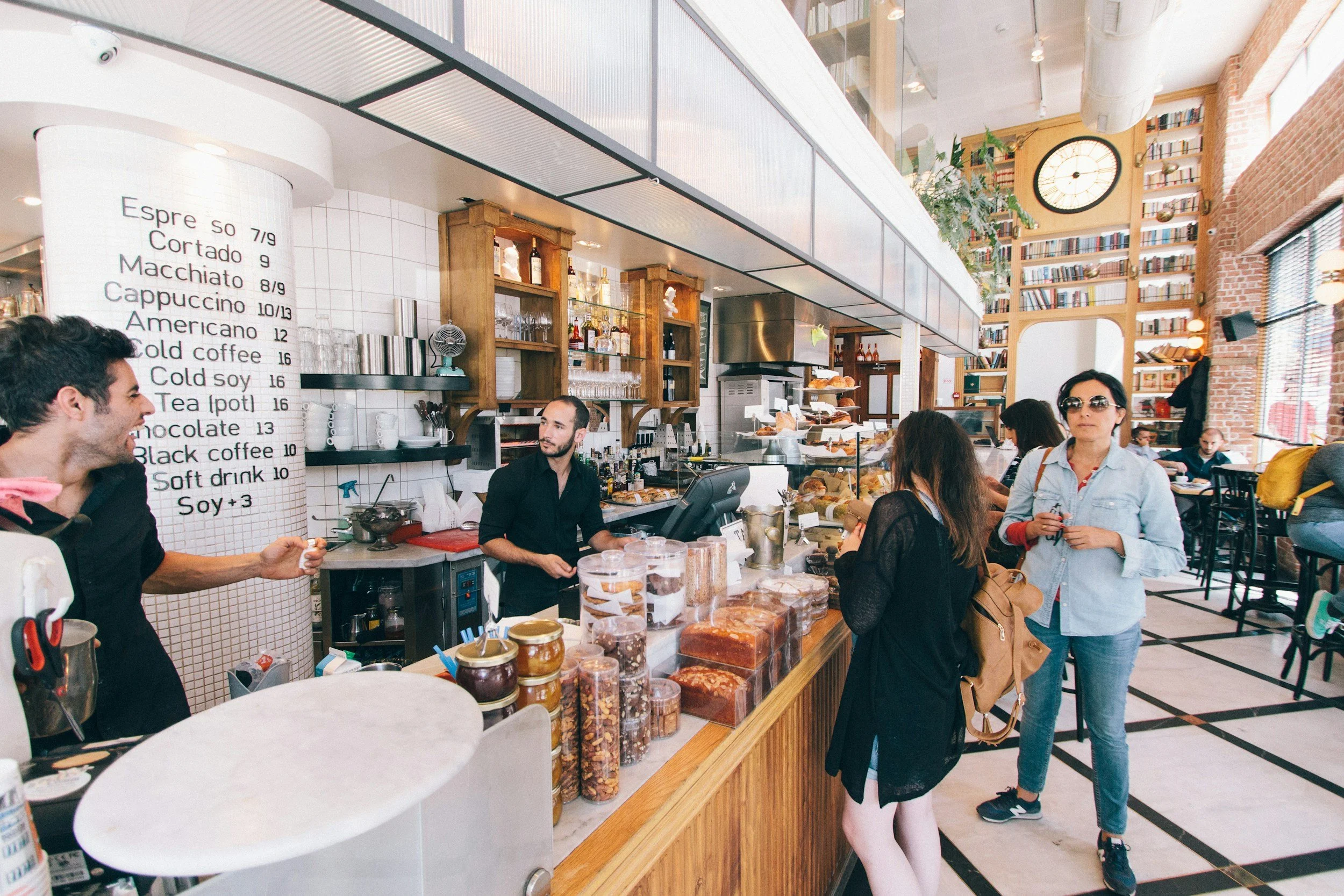 People ordering and chatting at a bright, modern coffee shop with a menu board, pastries, and a large clock on the wall.
