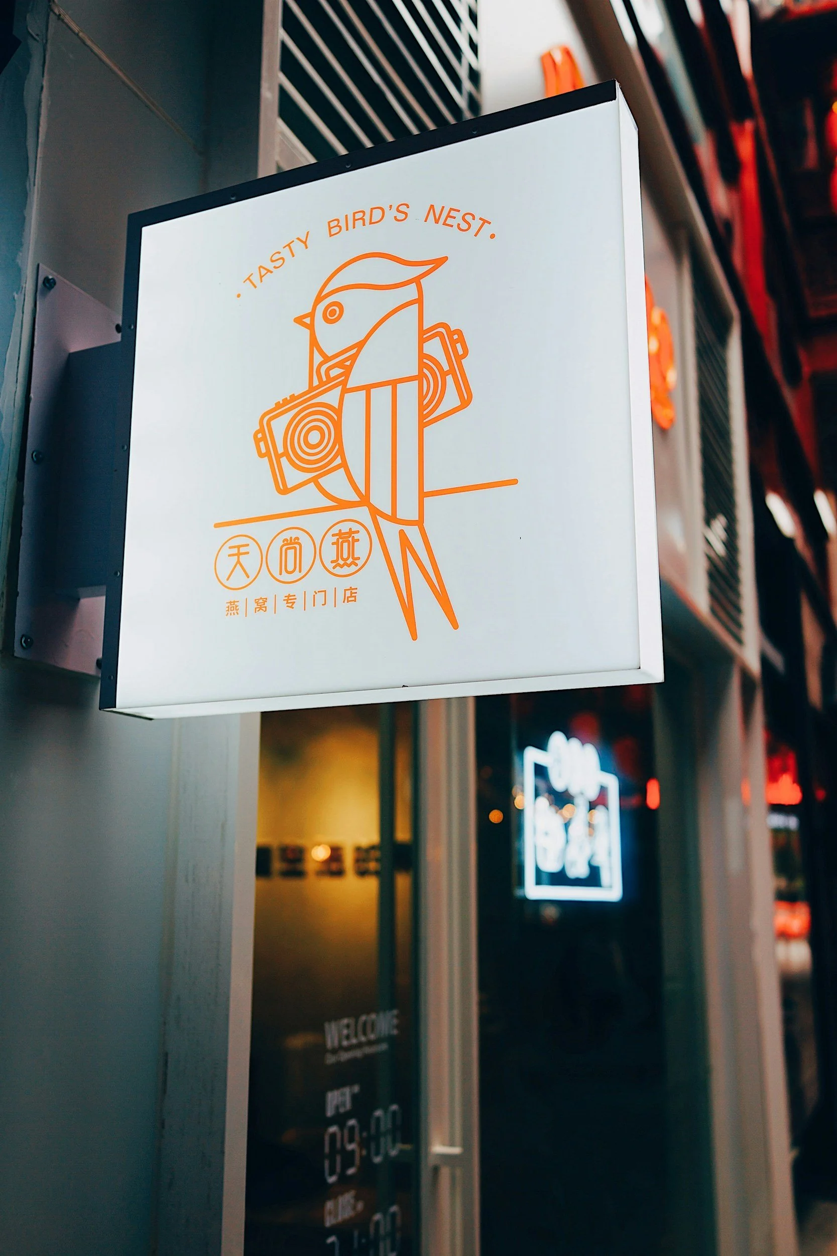 An illuminated sign for a restaurant called 'Tasty Bird's Nest' features a line art illustration of a bird with a camera on a white background, with Chinese characters and English text.