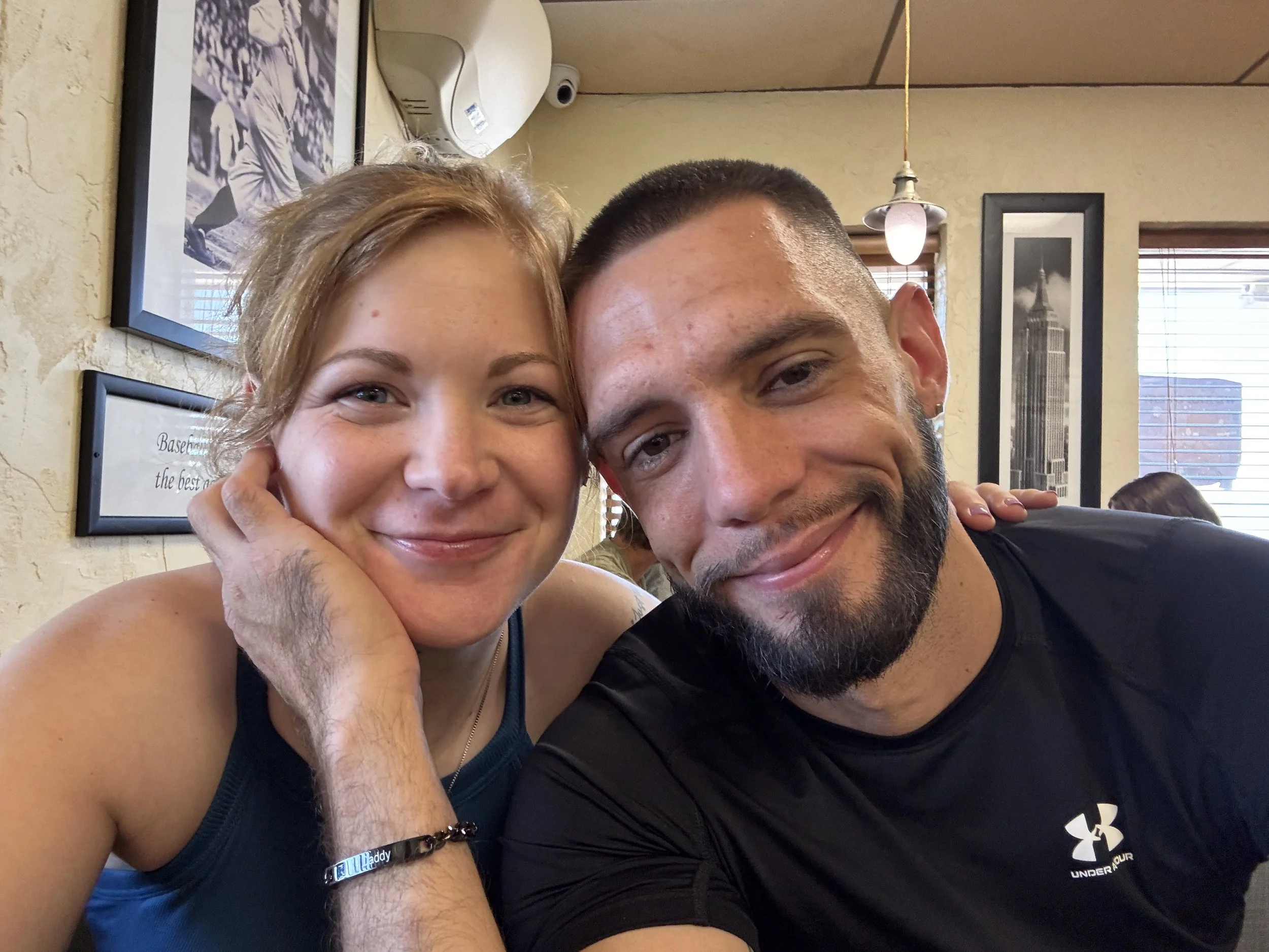 A young woman and a man smiling at the camera in a cafe or restaurant. The woman has red hair and is wearing a blue tank top. The man has short dark hair, a beard, and is wearing a black Under Armour shirt. They are sitting close together, and the woman has her hand on her cheek. The background shows framed pictures, a window, and a hanging light.