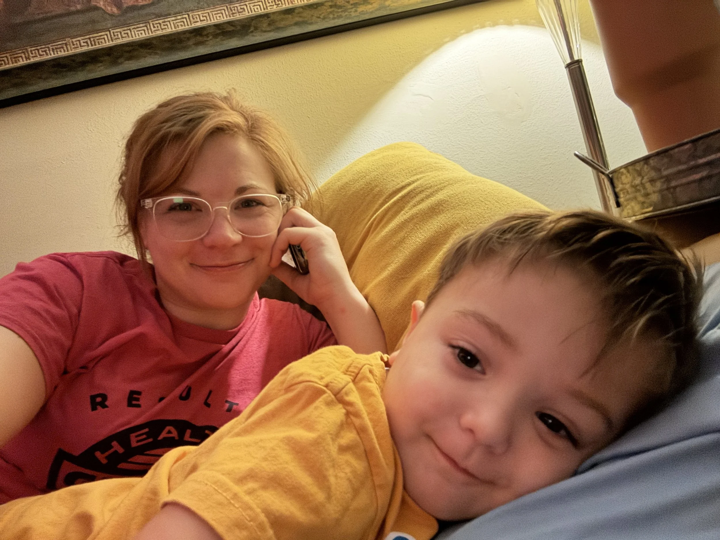 A woman with short red hair, glasses, and a pink T-shirt is smiling and talking on her phone, lying on a yellow couch. A young boy with short brown hair, wearing a yellow shirt, is lying next to her and smiling, resting his head on a blue pillow.