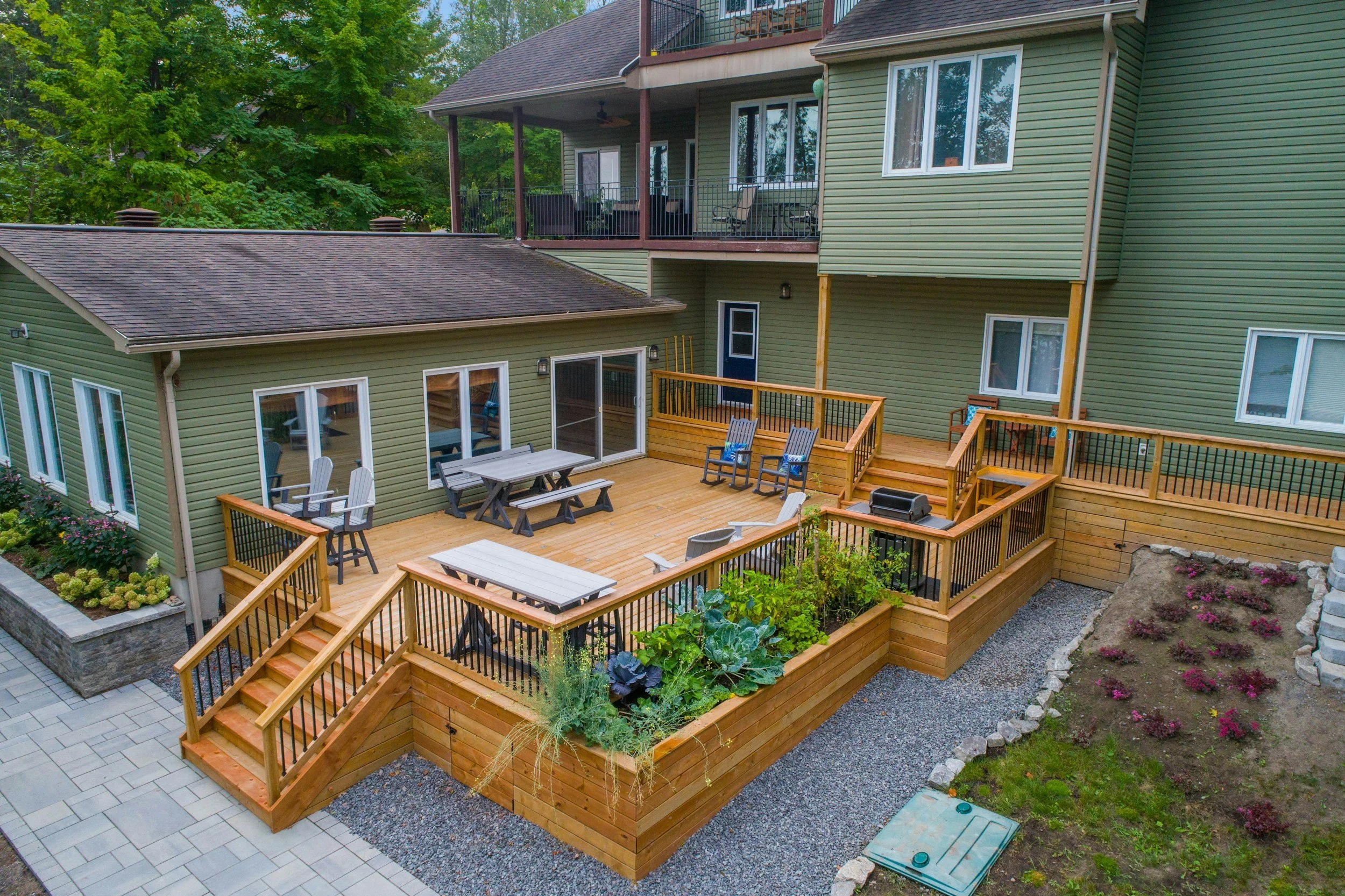 Backyard with wooden deck, outdoor dining table and chairs, potted plants, stairs, garden with flowers and rocks, patio area, and a green house with multiple windows and balconies at the upper level, surrounded by trees.