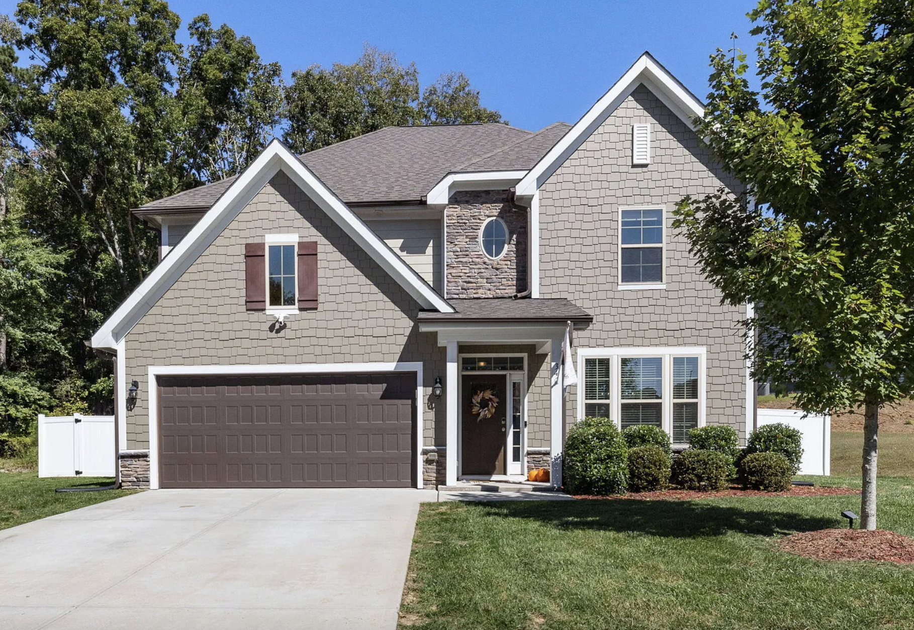 Front view of a two-story house with beige siding, brown shutters, a brown garage door, and a small front porch decorated with a wreath. There are three bushes in front and a tree to the right, with a blue sky in the background.