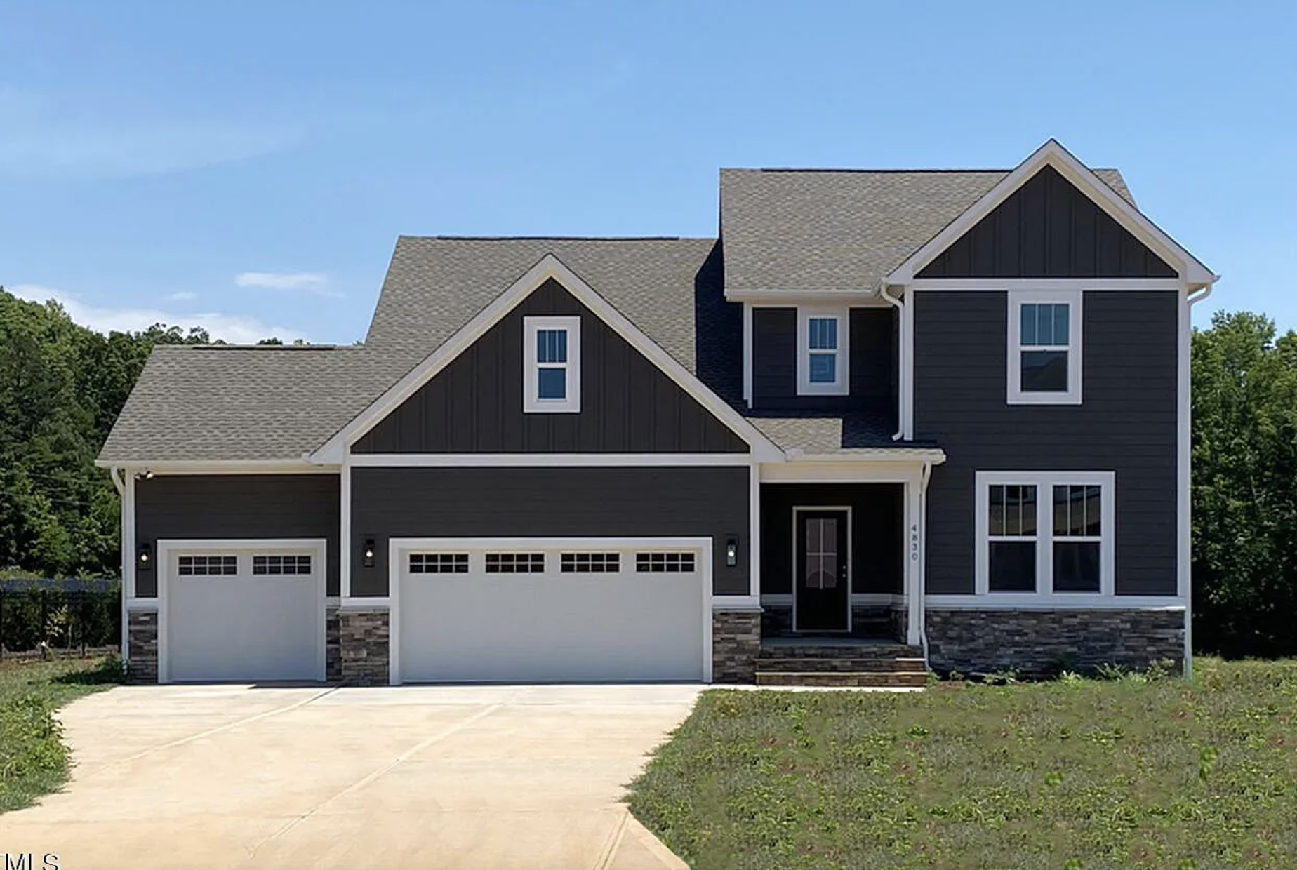 A modern two-story house with dark gray siding, white trim, and a stone accent on the lower exterior. It features a double garage doors, multiple windows, and a front porch with steps leading up to the black door. The house is surrounded by a driveway and some greenery, with trees in the background under a clear blue sky.
