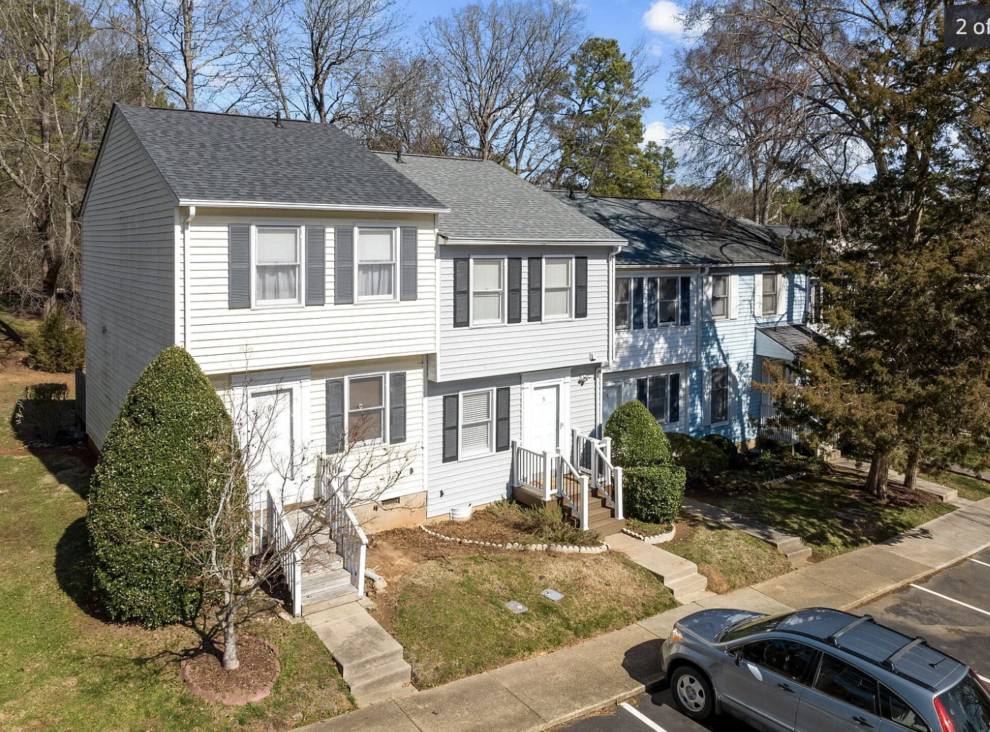 Multiple attached townhouses with white and blue siding, black shutters, and small front porches, surrounded by trees and parked cars.