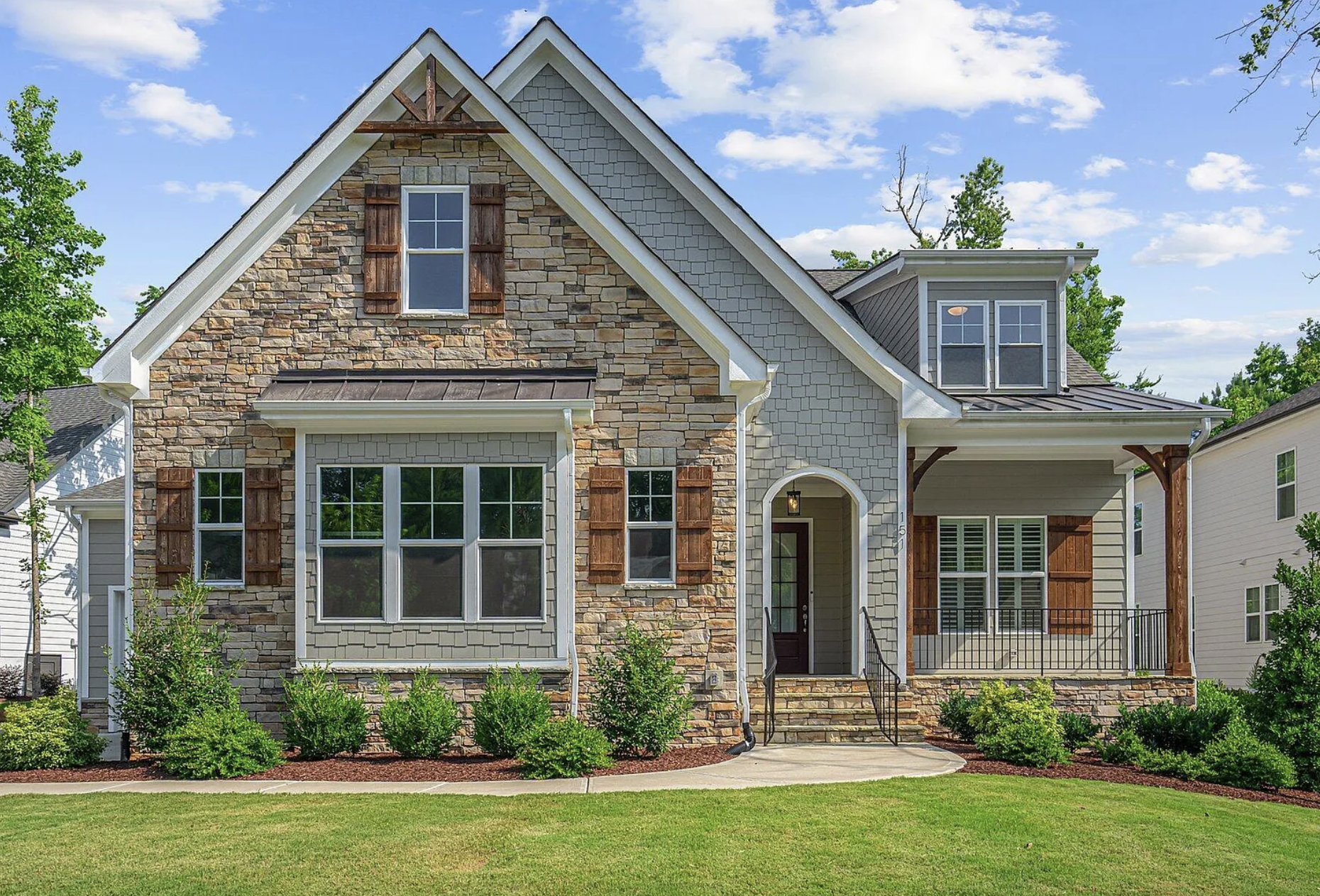 Front view of a two-story house with brick and beige siding exterior, large front window, small trees and bushes, and a well-manicured lawn under a partly cloudy sky.