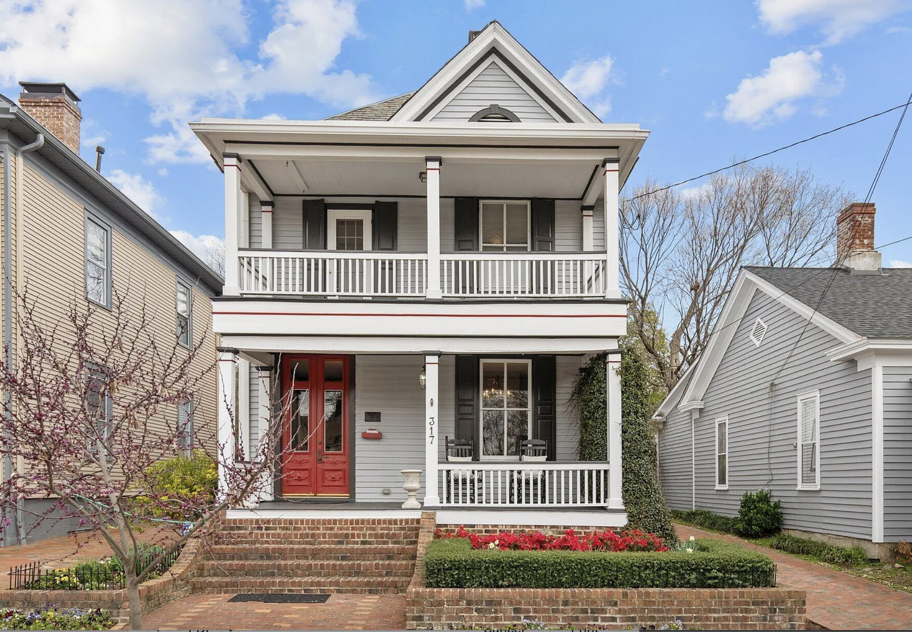 A two-story, gray wooden house with a front porch, white railings, and black shutters on the windows. The house has a red front door, steps with brick edging, a small front garden with a tree and colorful flowers, and neighboring houses on either side. The sky is partly cloudy with blue patches.