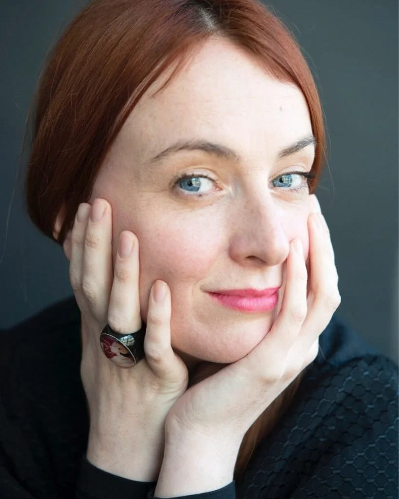 A woman with red hair and blue eyes looking at the camera and smiling with her head turned and hands on her face.
