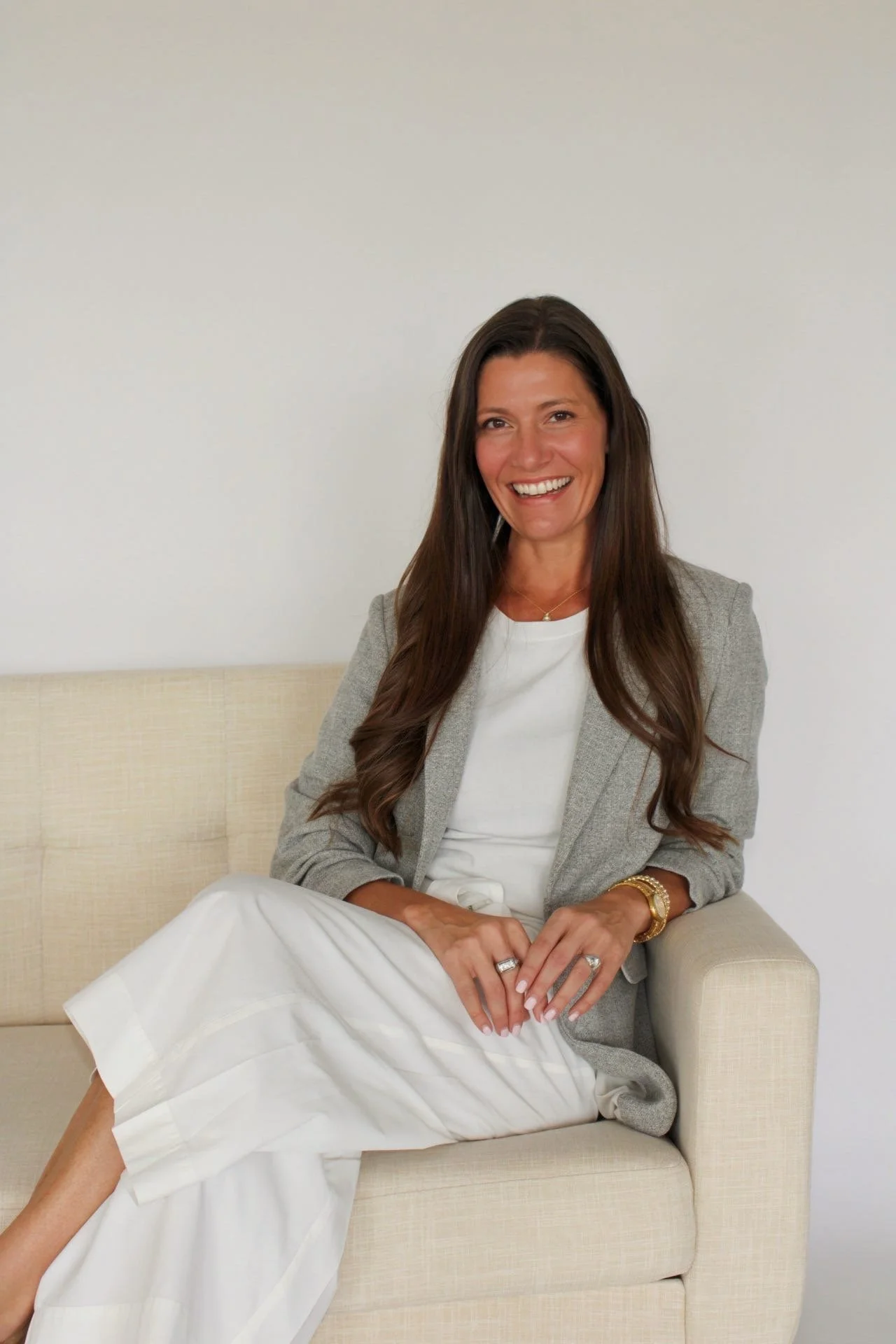 A woman with long brown hair sitting on a beige sofa against a plain white wall, smiling at the camera. She is dressed in a light gray blazer over a white top and white pants, with gold jewelry including rings, a bracelet, and a necklace.