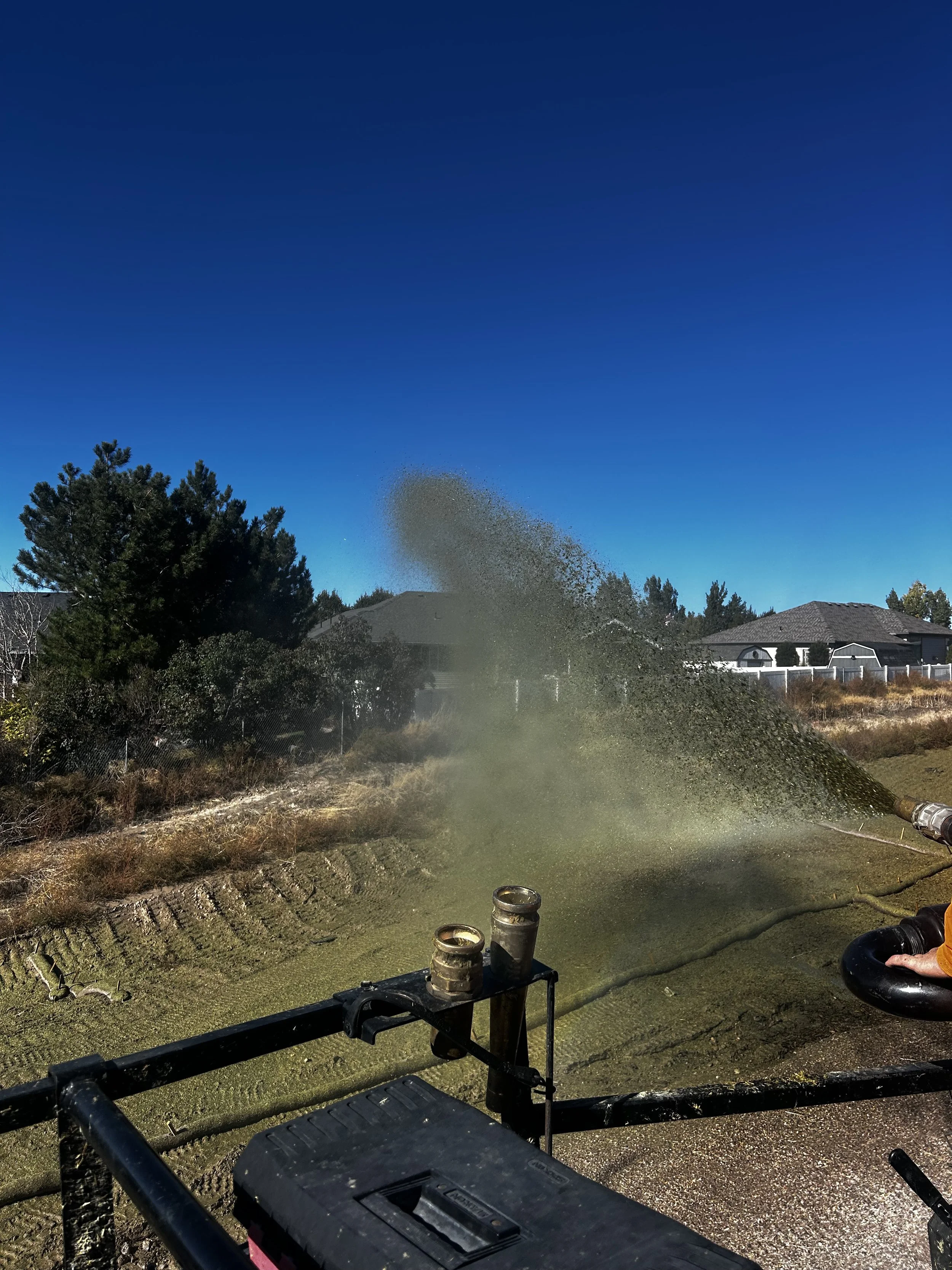 A hose spraying water onto the ground with a blue sky and houses in the background.