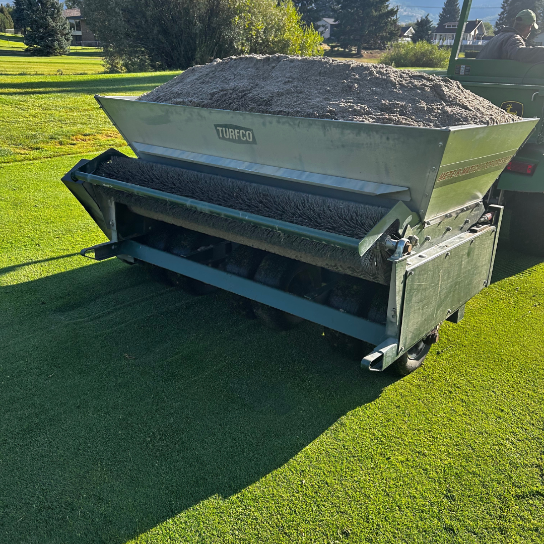 A turf aerator machine on a golf course with a large pile of sand or soil in the hopper.