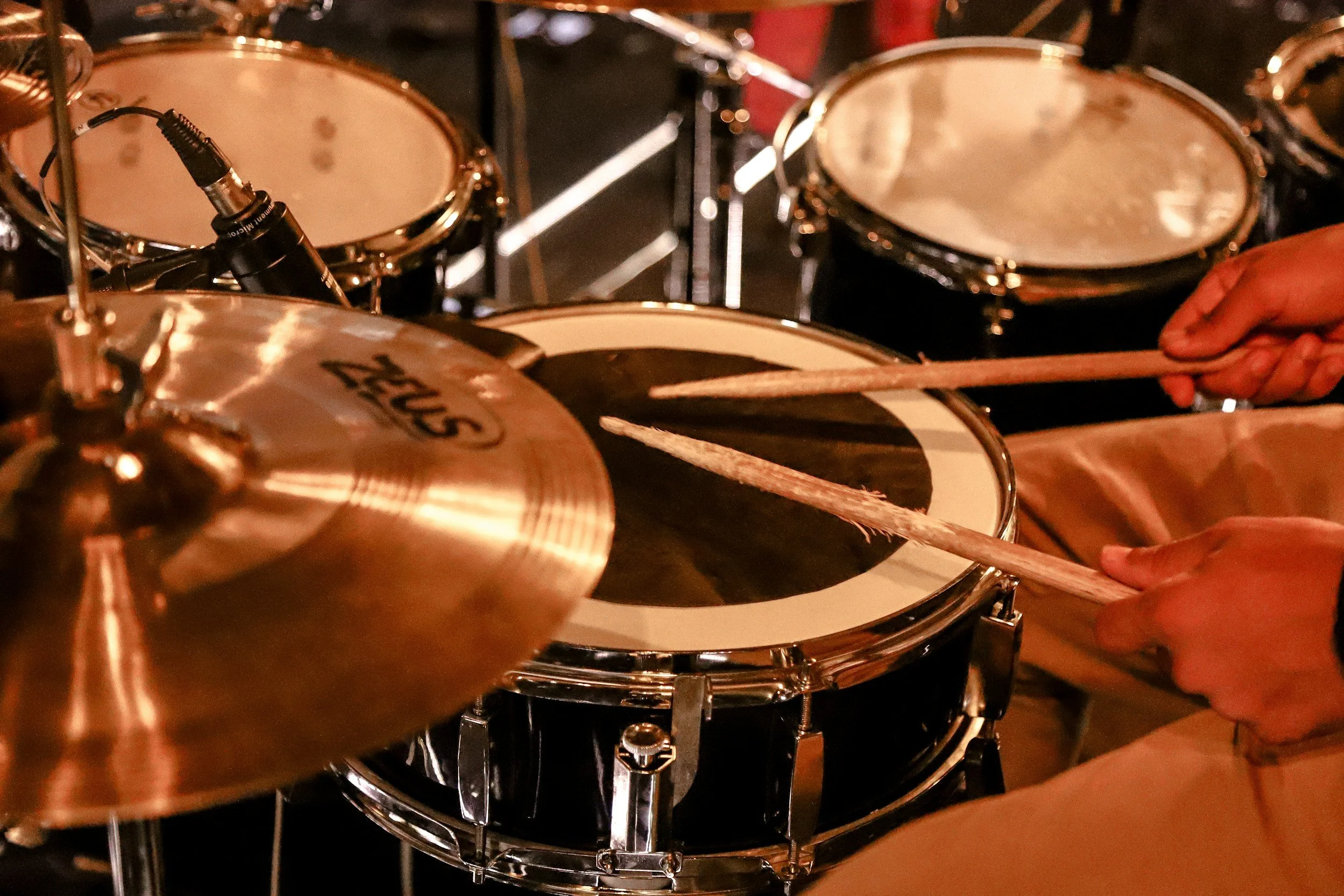 Close-up of a person playing a drum set, with hands holding drumsticks hitting a snare drum. Surrounding drums and cymbal are also visible.