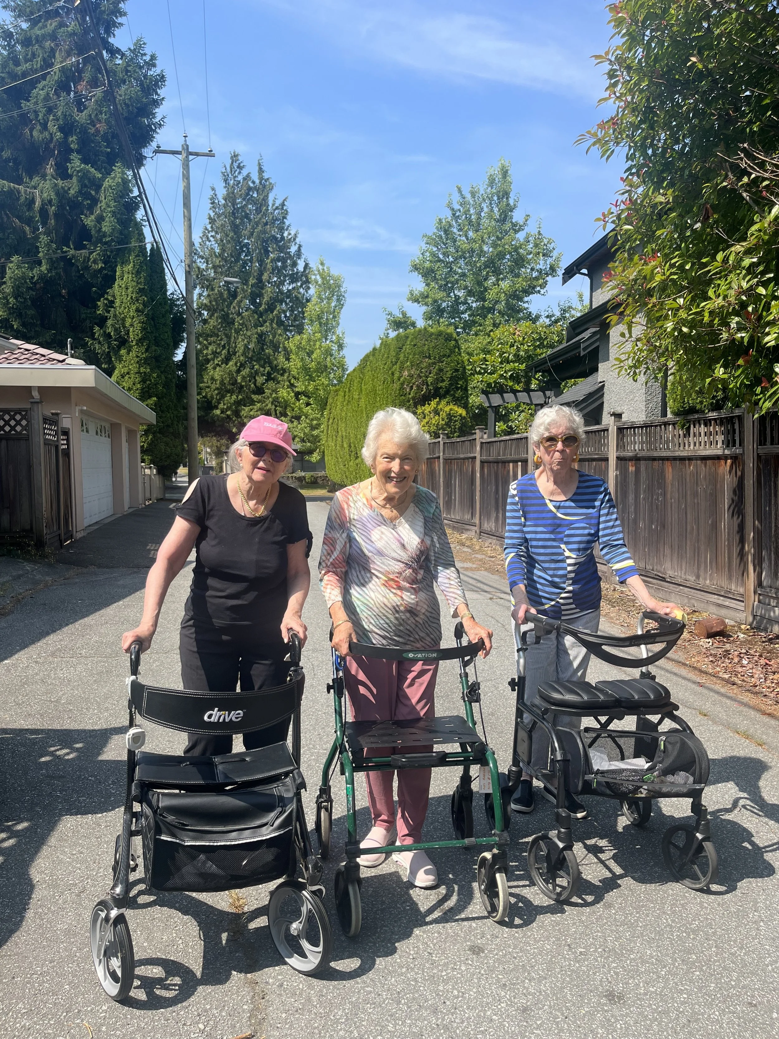 Three elderly women walking with assistive devices on a sunny street, surrounded by trees and houses.