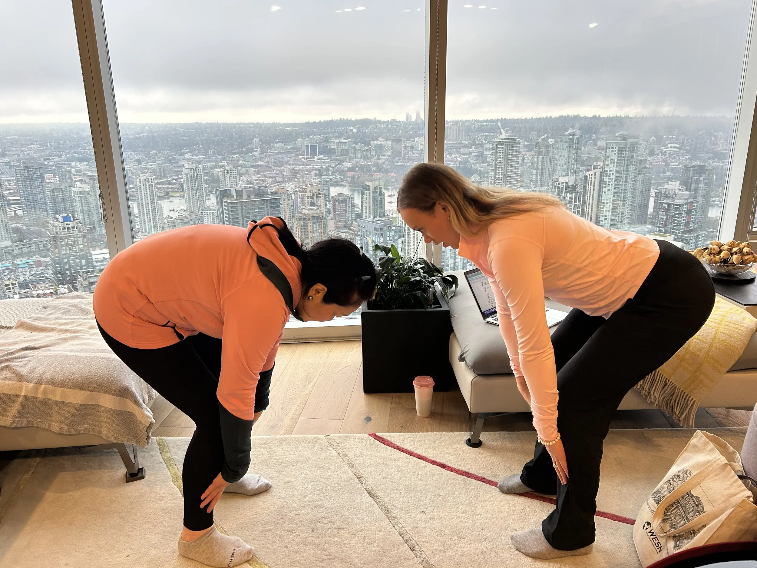 Two women are bending forward and touching their toes in a high-rise apartment with large floor-to-ceiling windows revealing a city skyline. One woman has dark hair, and the other has light brown hair. Both are dressed in light-colored shirts and dar