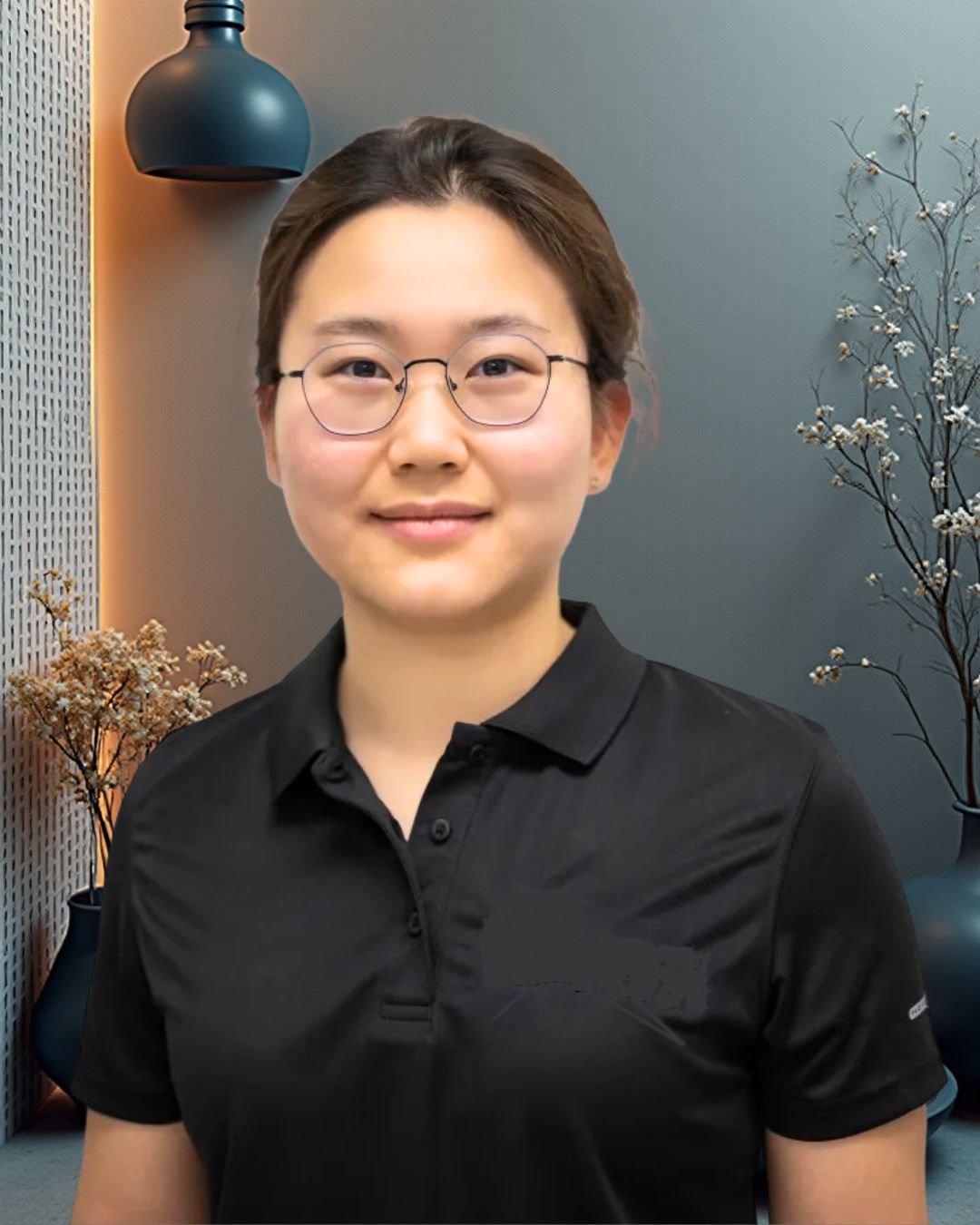 A woman with short brown hair and glasses smiling at the camera, wearing a black polo shirt, standing in front of a modern indoor wall with decorative vases and dried flowers.