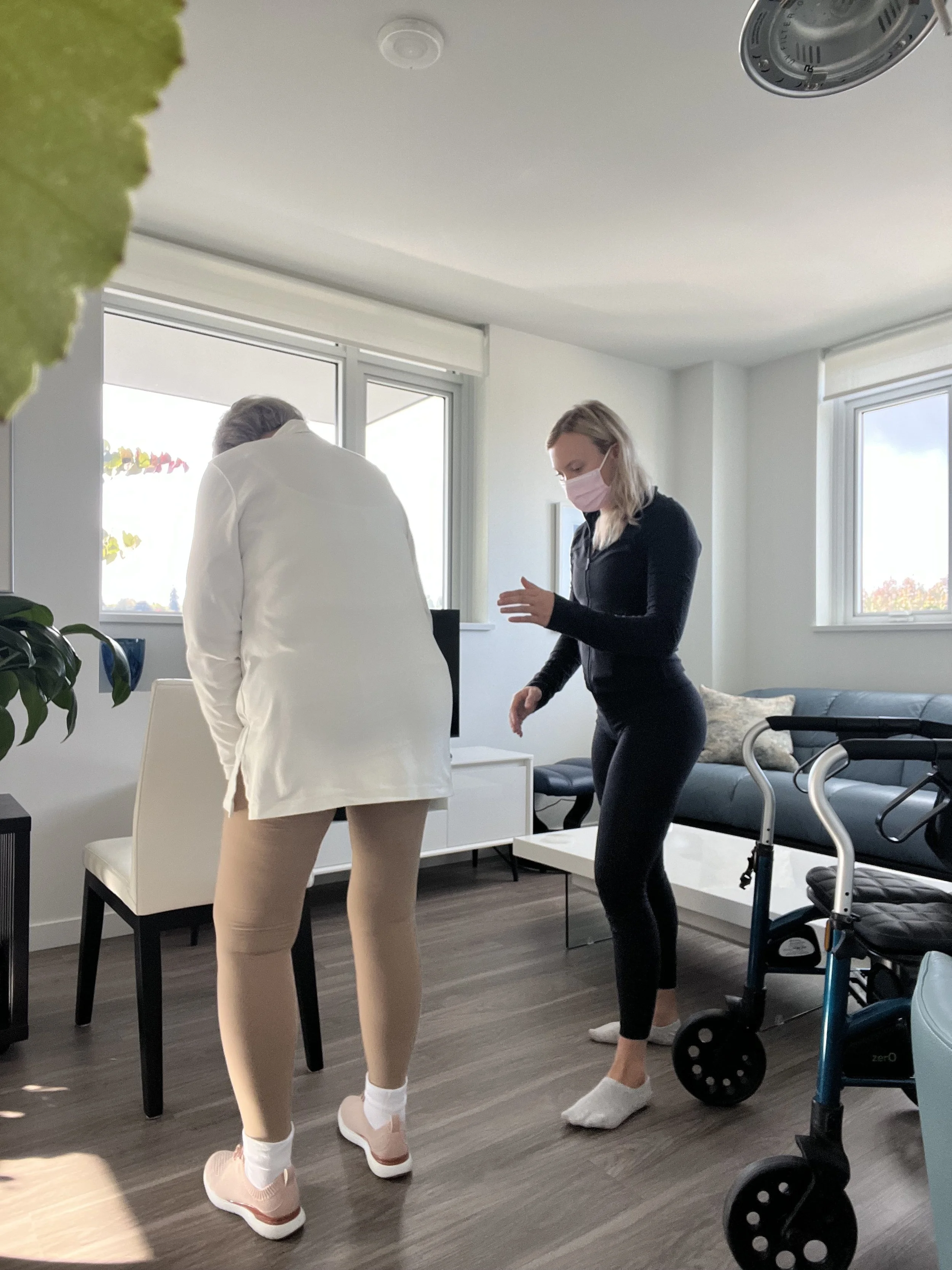 Two women, one in a white coat and the other in black, are in a bright room with large windows, wearing face masks. One is leaning over a table and the other is standing and gesturing.