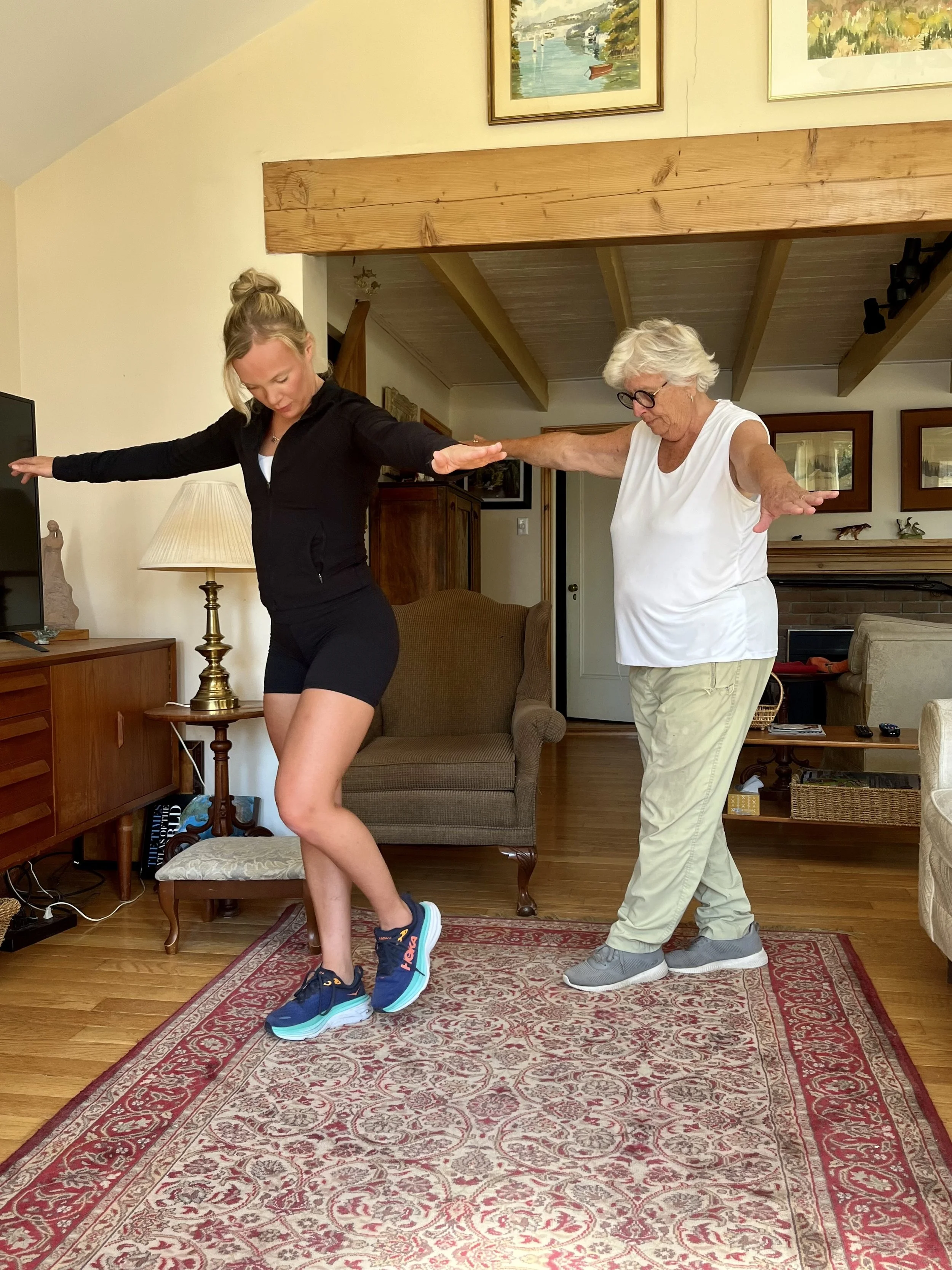 A young woman and an elderly woman practicing dance or exercise indoors. The young woman is striking a pose with her arms outstretched, wearing athletic clothes and sneakers. The elderly woman is behind her, mimicking the pose, wearing glasses, a whi