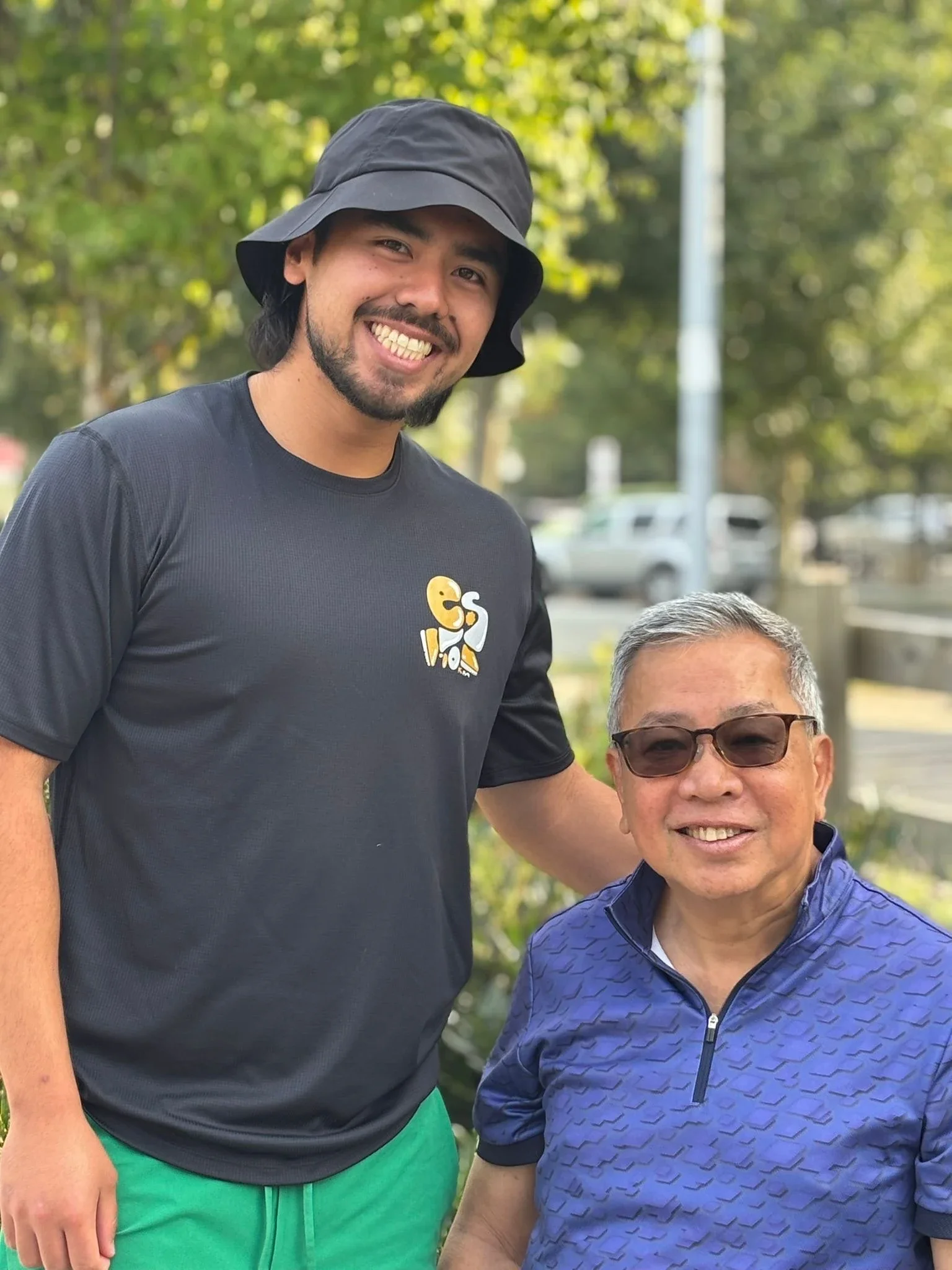 A young man wearing a black bucket hat and a black T-shirt with a yellow and white graphic. An older man with glasses and a blue patterned jacket. They are outdoors with trees and parked cars in the background, smiling.