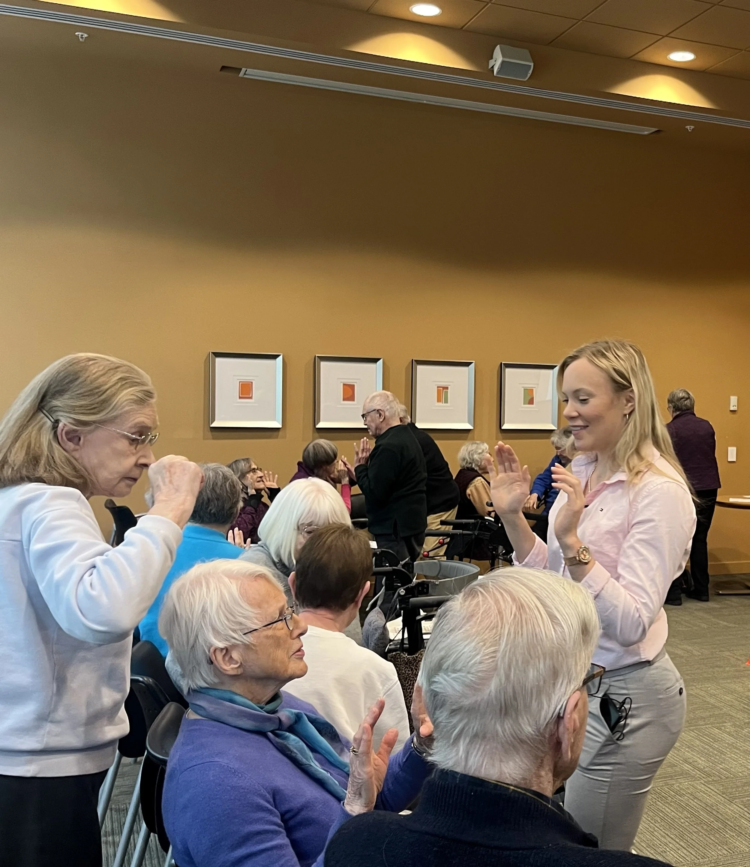 A group of elderly and middle-aged people gathered at Homestretch Brain & Balance program in Seasons retirement home. They are engaging in conversations, with some standing and others seated, and appear to be enjoying themselves.