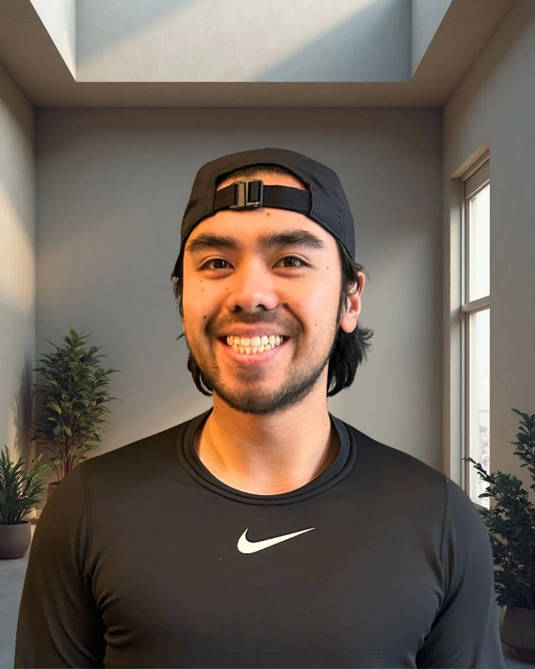 Smiling young man wearing a black Nike athletic shirt and a backward black cap, standing in a room with potted plants and a window, with a skylight in the ceiling.