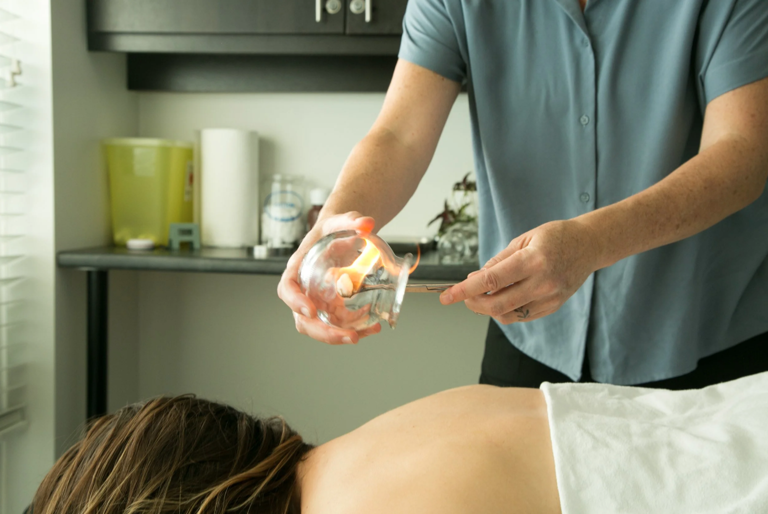 A person is pouring a small flame from a glass Incense holder onto a rice grain during an aromatherapy or healing treatment, with a client lying face down on a massage table.