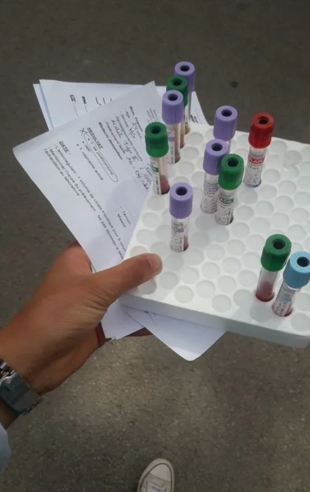 Hand holding a white tray with blood sample vials and printed papers over a concrete surface.