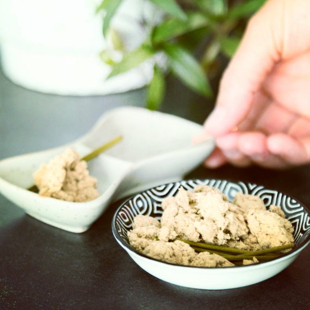 A hand holding a spoon over a bowl of beige crumble or powder, with a small dish containing the same substance in the background on a black surface, and a green plant in the blurred background.