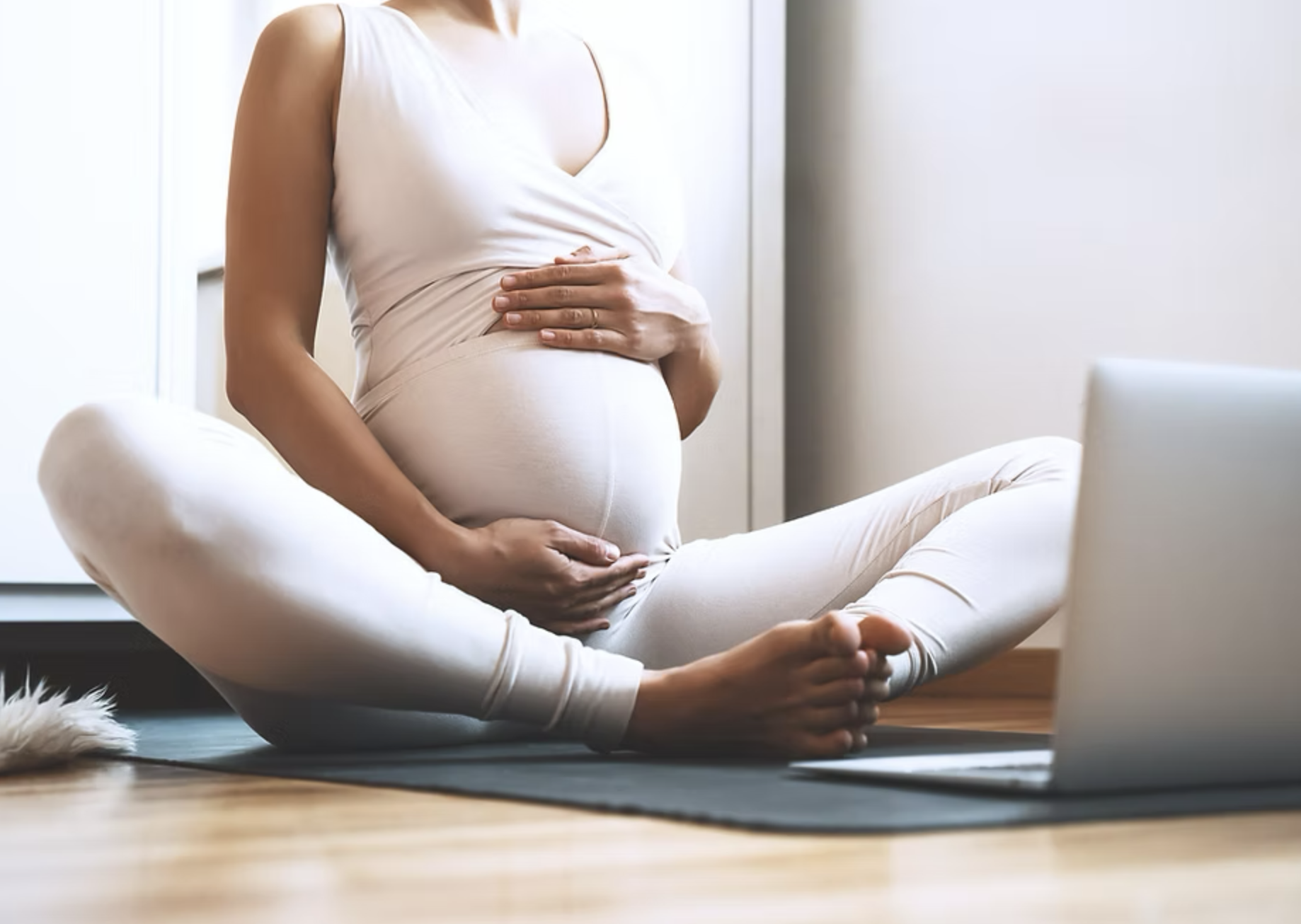 A pregnant woman practicing yoga indoors, sitting cross-legged on a yoga mat while looking at a laptop.