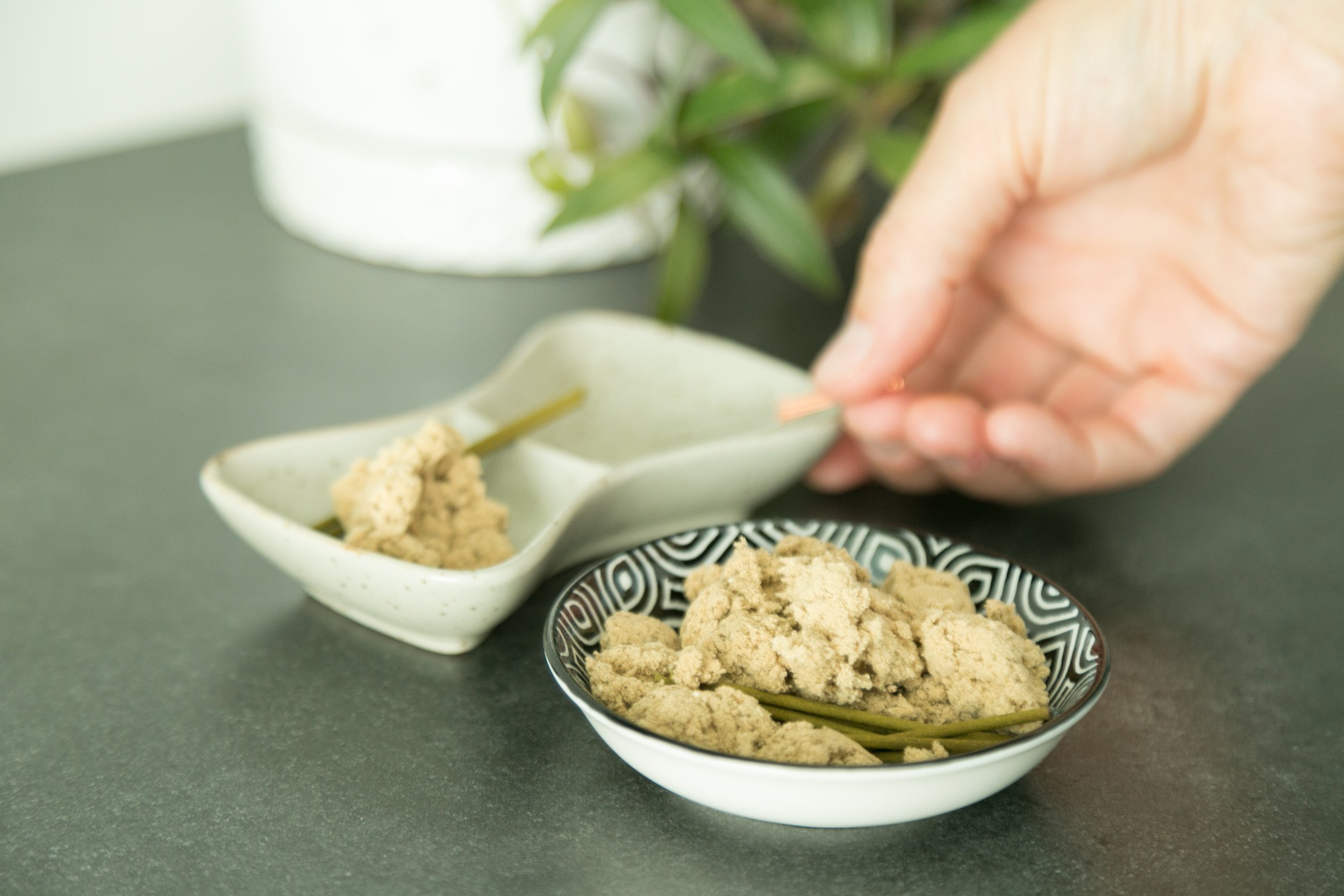Hand sprinkling powdery substance from a white and black patterned bowl into a small rectangular dish with similar powder. The background features a black surface and a blurred green plant.