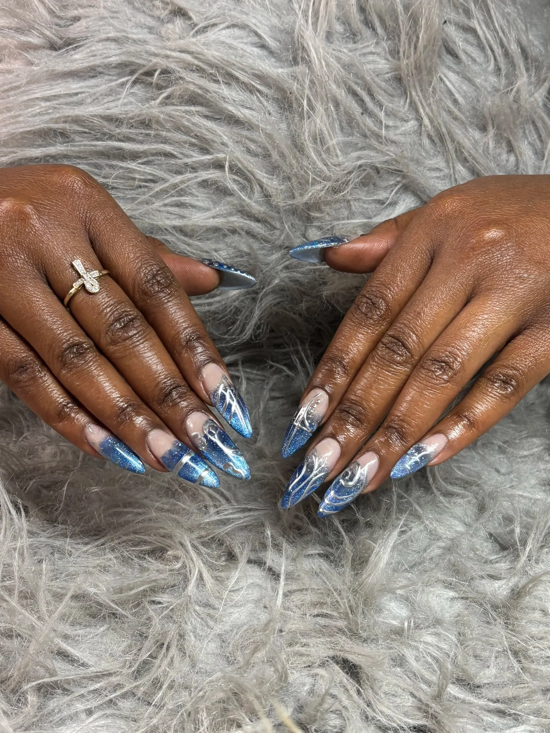 Close-up of hands with shiny blue and white nail art, resting on a fluffy grey surface, with a ring on the left ring finger.