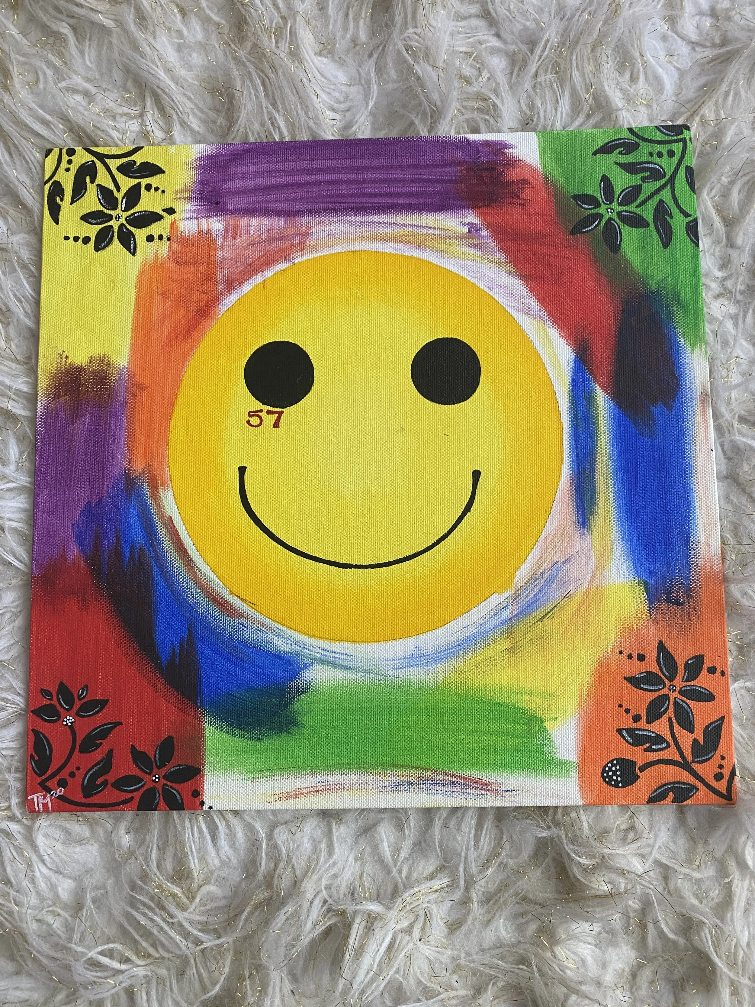 Colorful painting of a large yellow smiley face in the center, surrounded by multicolored brushstrokes and black floral designs in each corner.