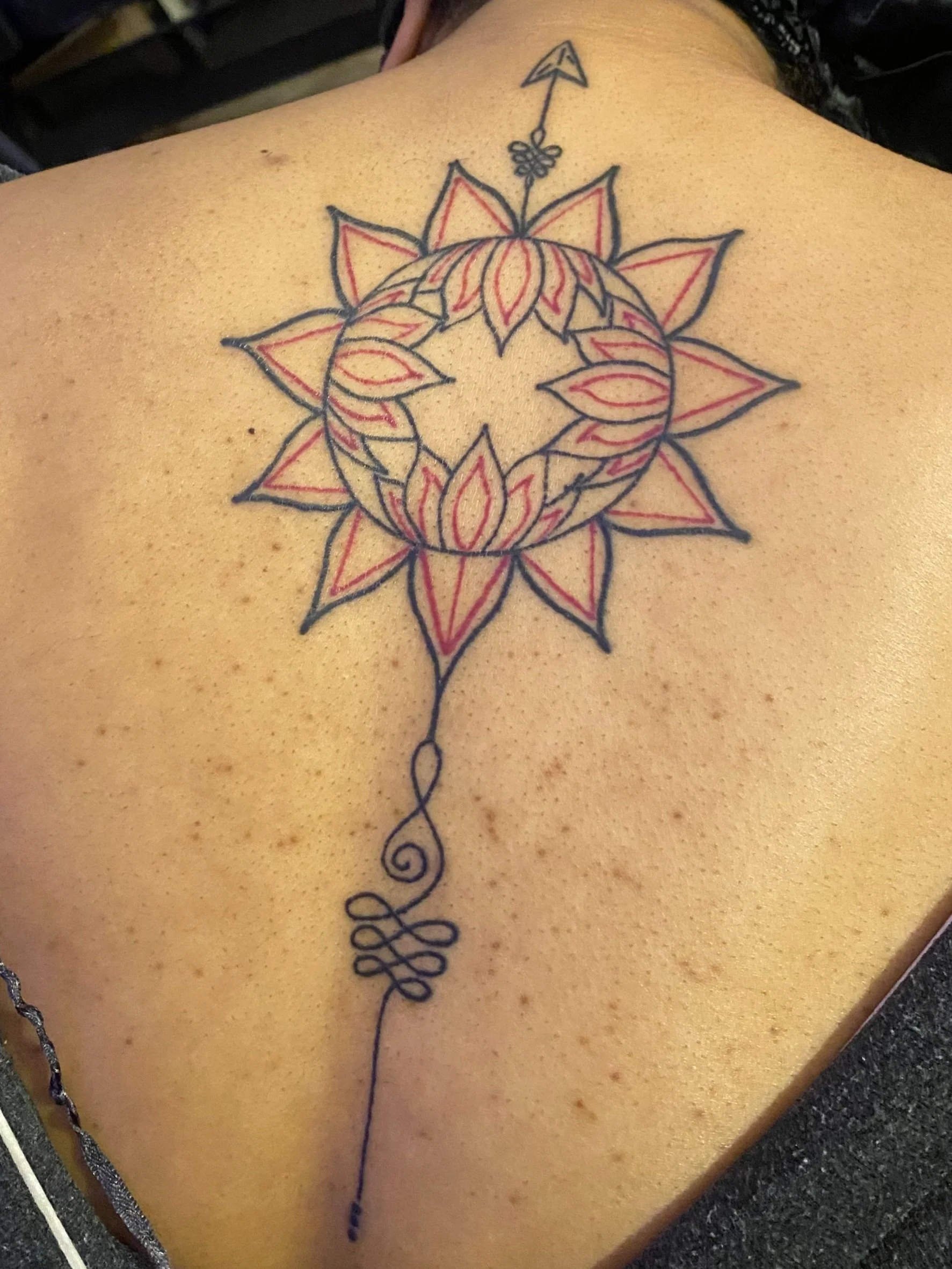 A fresh tattoo of a geometric sun design with an arrow and a decorative string hanging down, located on a person's upper back.