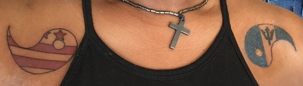 Two tattoos on shoulders, one of a crescent moon with stripes and a star, and the other of a circular symbol with a V-shape, an eye, and wings.