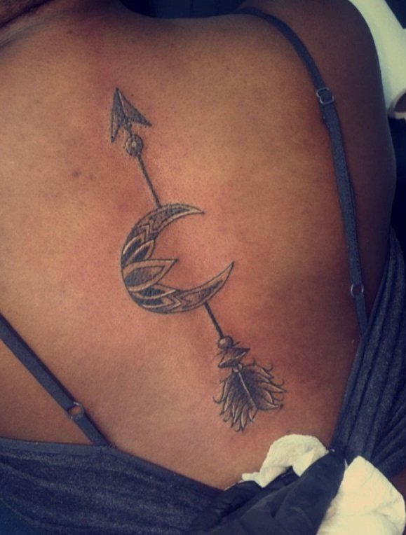 Tattoo of a crescent moon with tribal patterns, an arrow passing through it, and a feather hanging from the arrow on a person's shoulder.