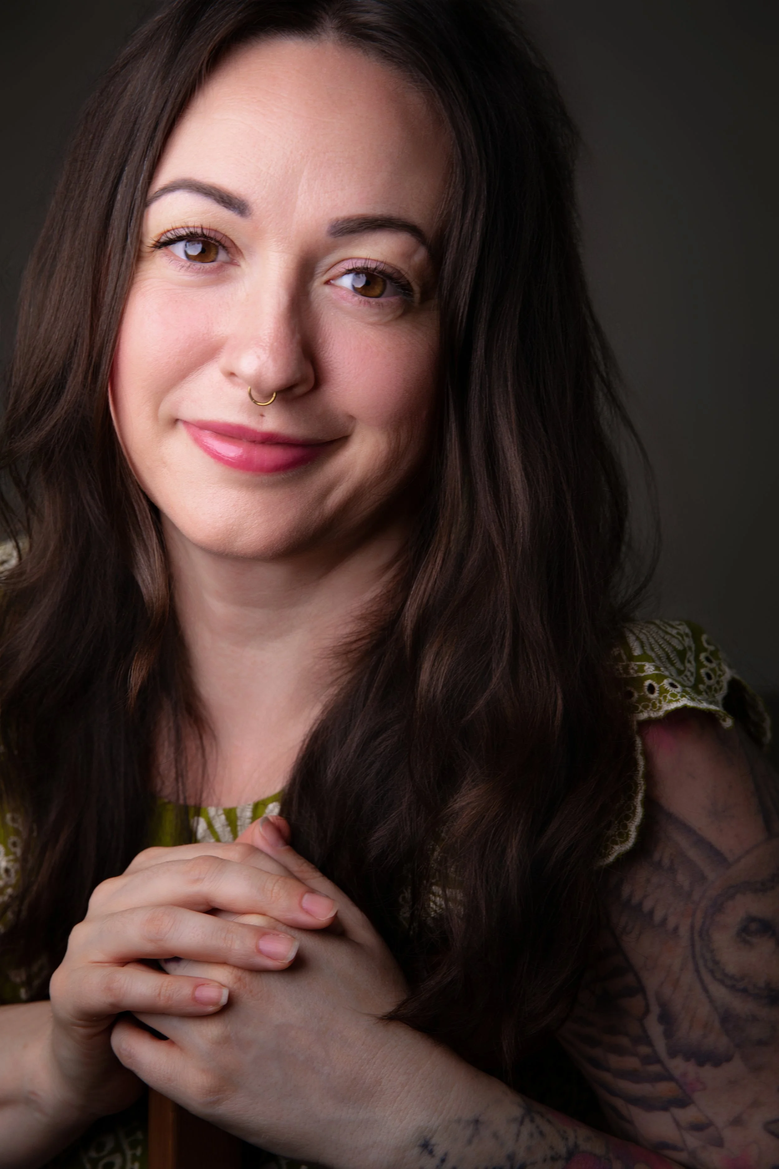 A woman with long dark hair, brown eyes, and a septum piercing smiles softly. She has tattoos on her arm and is holding her hands together in front of her. She is wearing a green patterned top, and the background is dark.