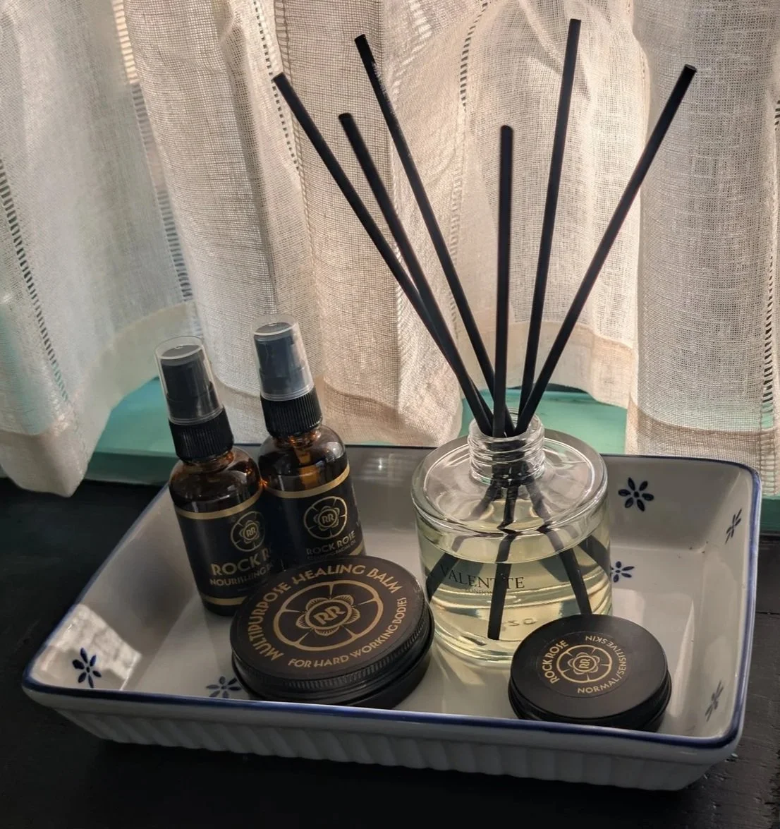 A tray containing two small spray bottles labelled 'Rock Rose' with black and gold labels, two small tins of 'Rock Rose' moisturizing and healing balm, and a glass diffuser with black reed sticks filled with a clear liquid.