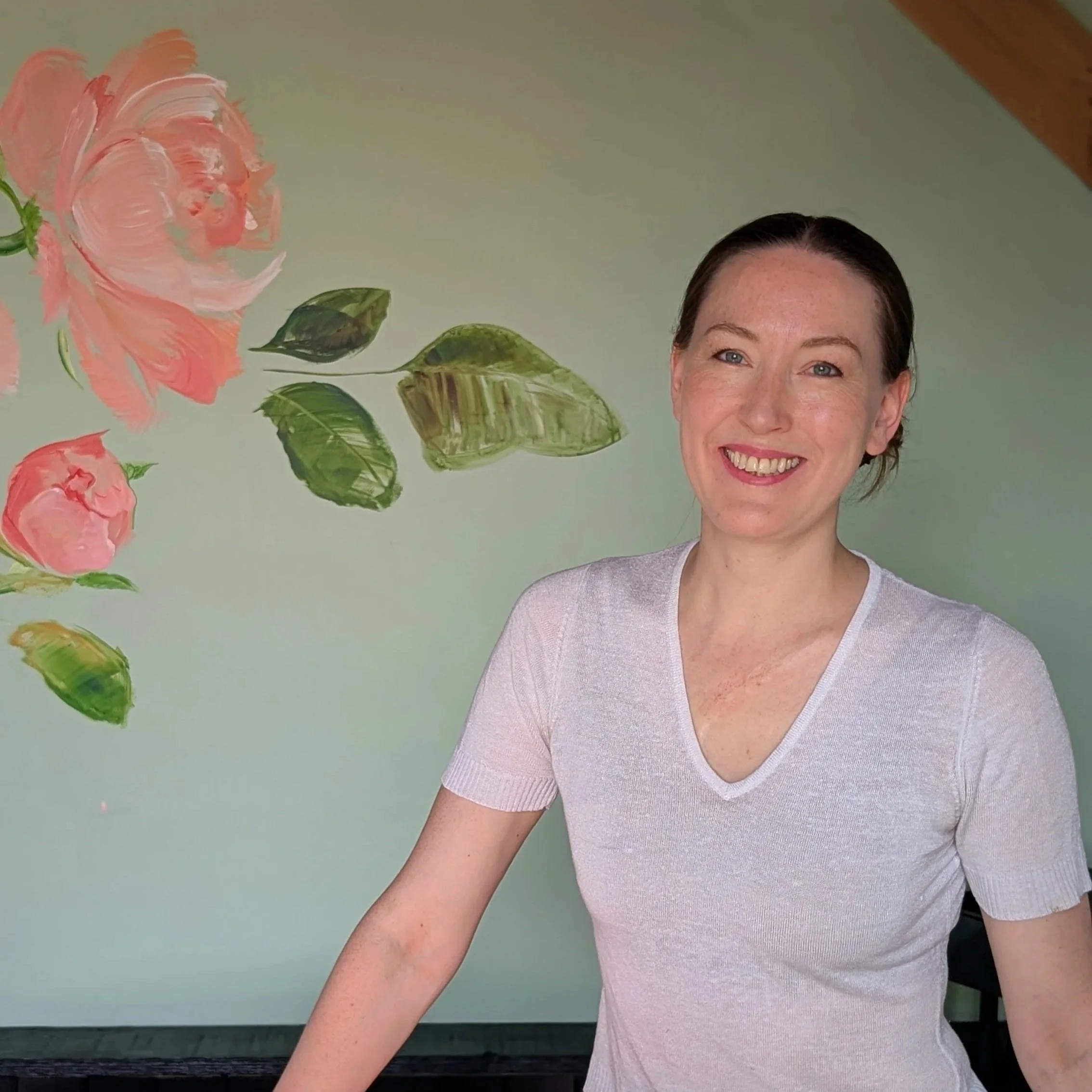 Rosie, smiling, wearing a white V-neck shirt, in front of a pale green wall with painted pink roses and green leaves.