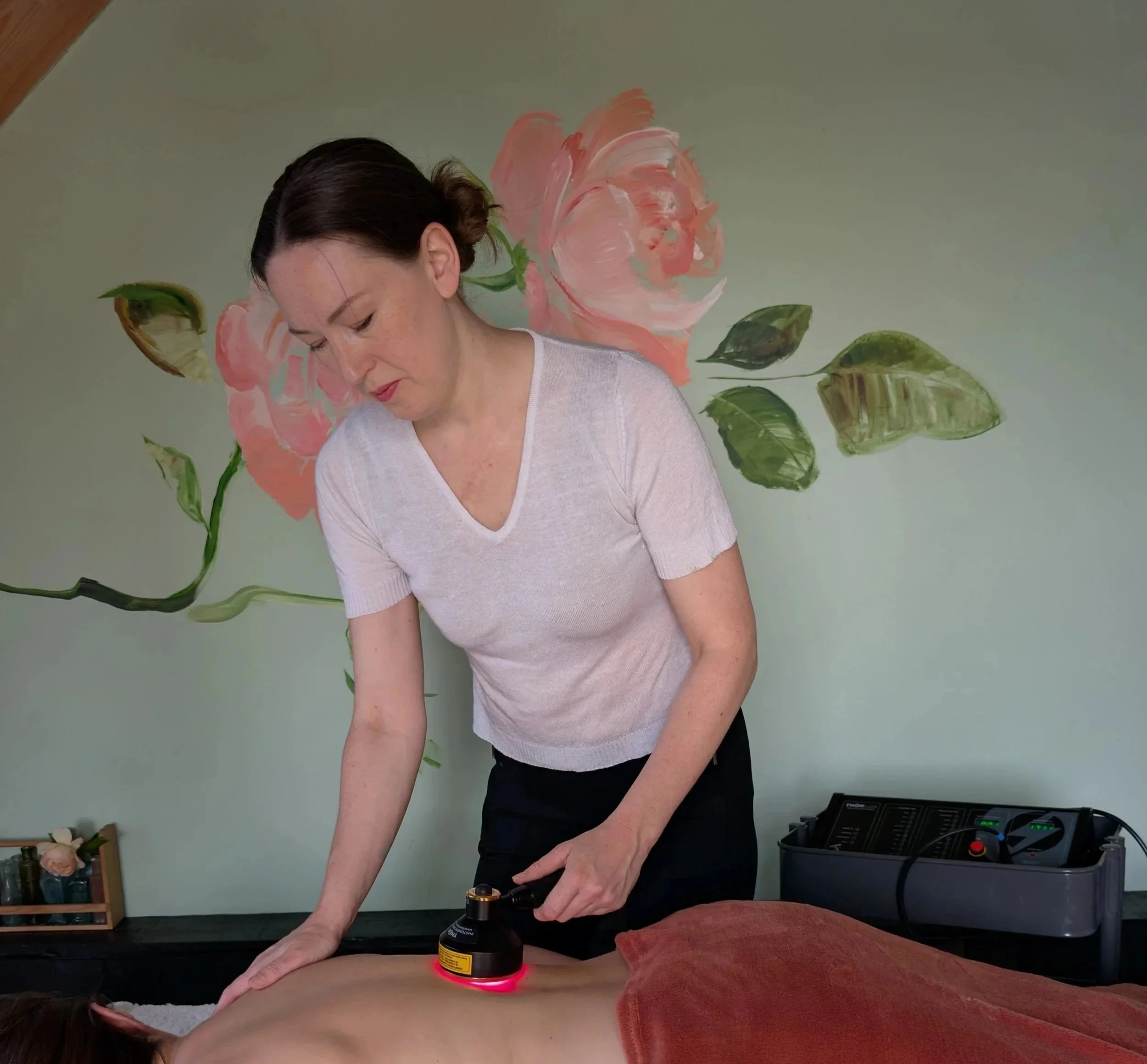 Rosie, with her hair tied back and wearing a white top, is performing red light therapy on a client. The background features a wall painted with large pink roses and green leaves. The red light therapy machine is at the back of the scene.