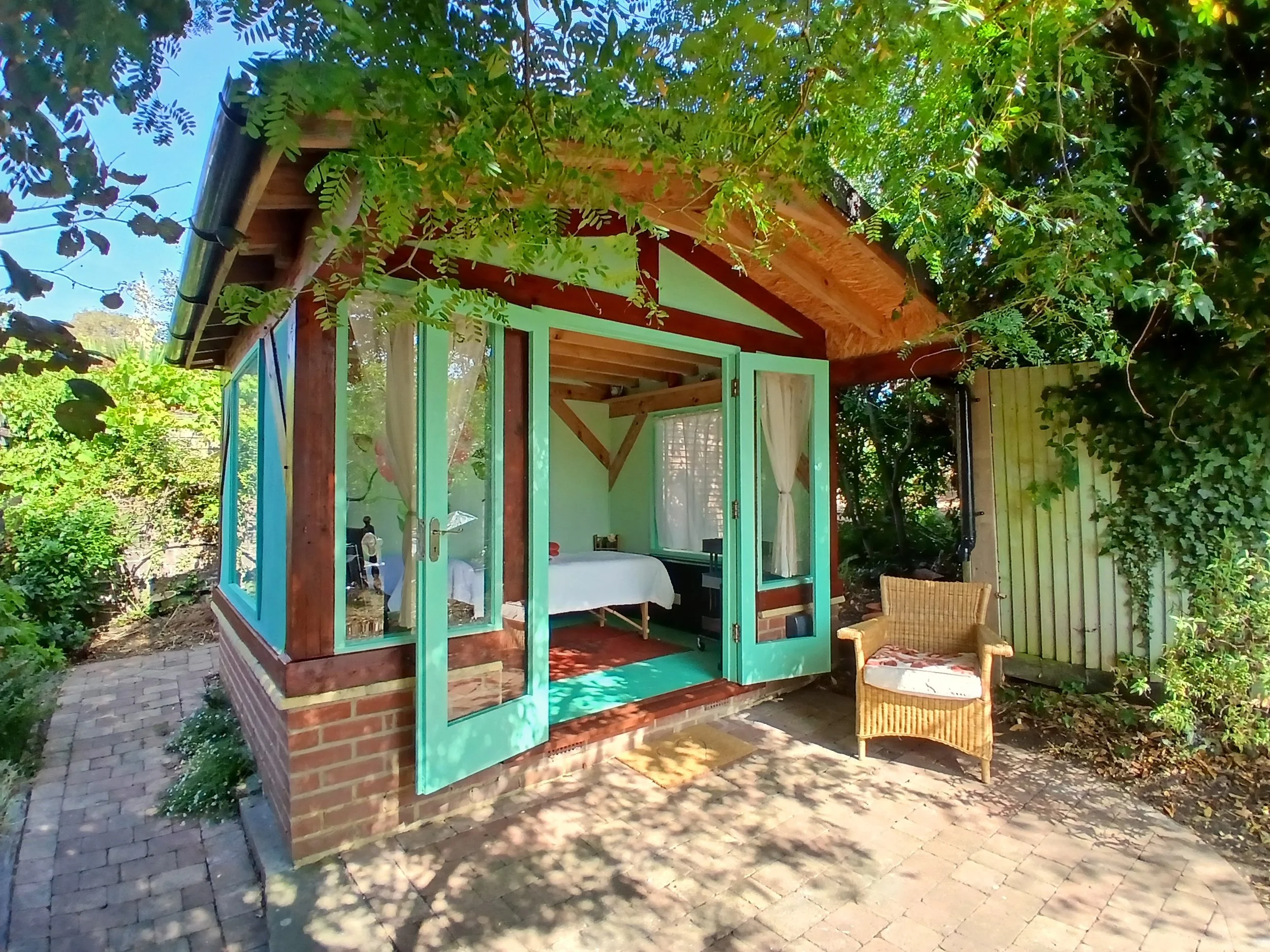External view of the Rose Clinic, with timber framing and green windows. The room has French doors that open to a patio with a wicker chair nearby. The area is surrounded by lush greenery and trees, providing shade and a tranquil atmosphere.