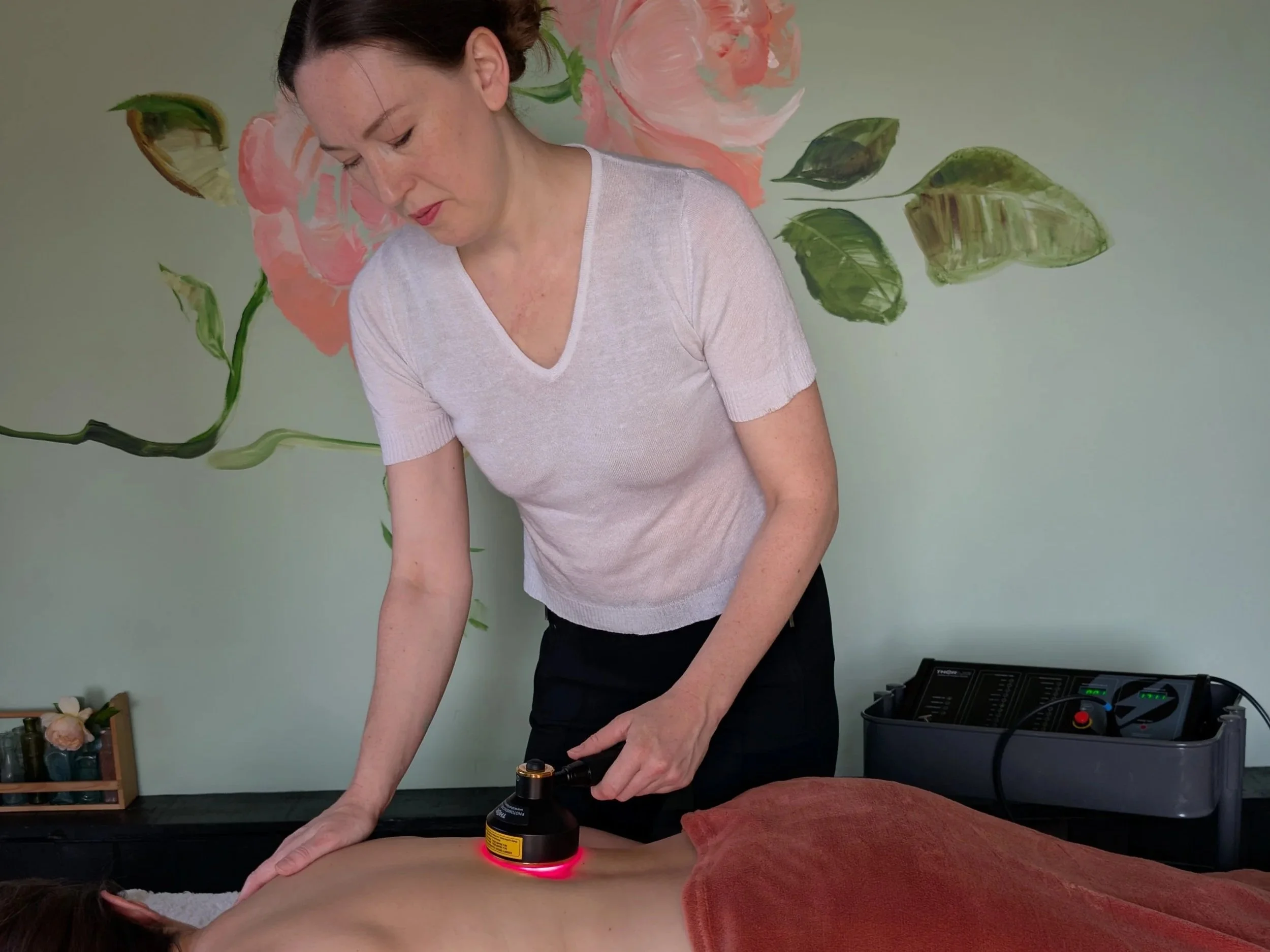 Rosie giving red light therapy treatment to a client, inside The Rose Clinic, a rose mural is visible on the pale green wall behind her. 