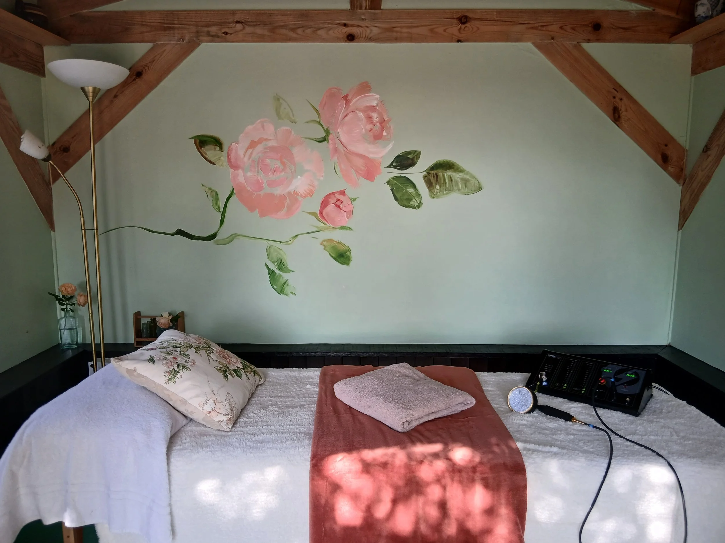 Interior of The Rose Clinic showing the treatment bed with cosy white and peach blankets, the red light therapy machine is visible, as are a beamed ceiling and pale green walls with a hand-painted rose mural.