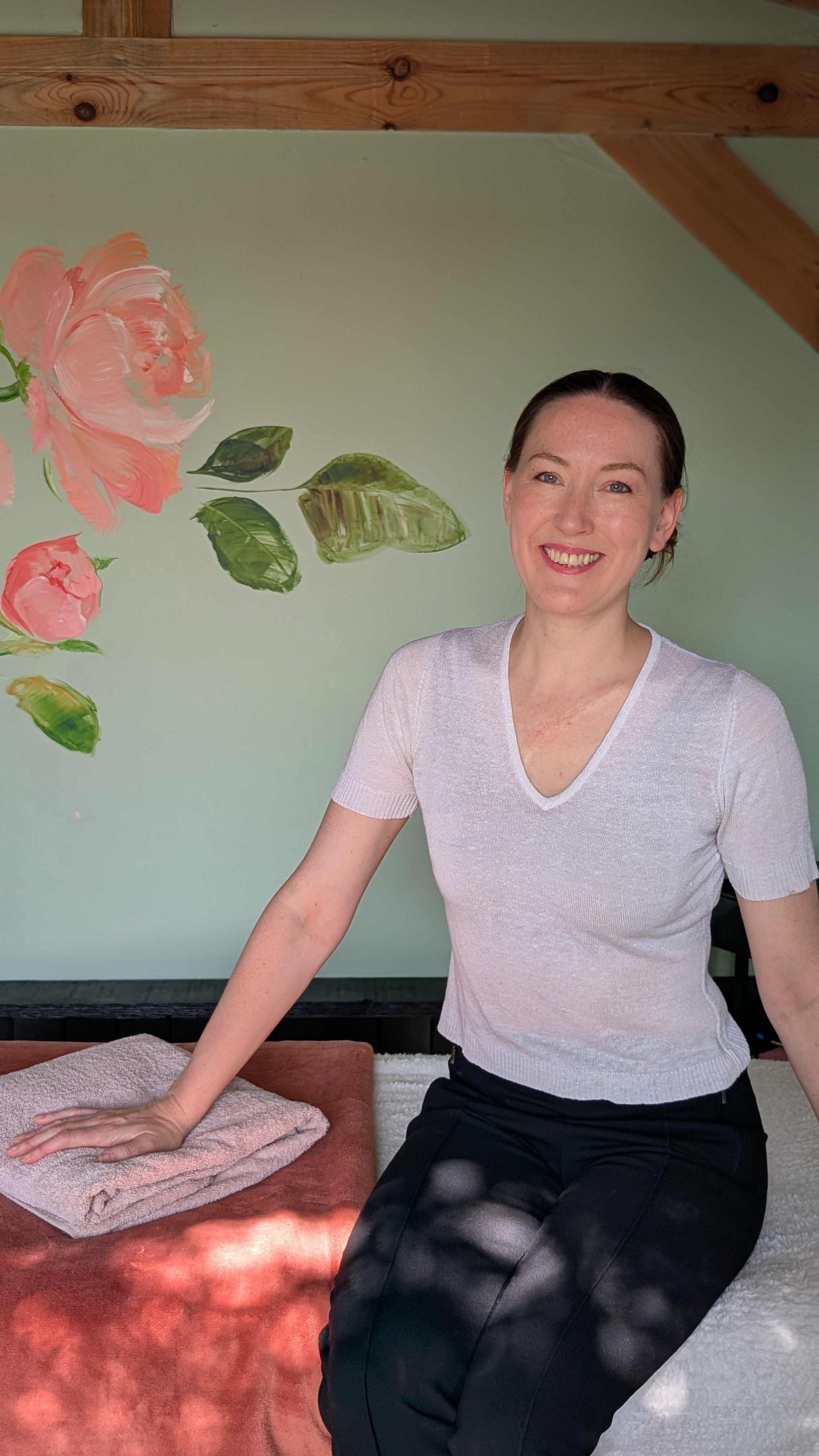 Rosie, smiling, wearing a light-colored v-neck shirt and black pants, leaning on the treatment table with a folded pink towel. Behind her is a wall painted with pink roses and green leaves, and part of a wooden ceiling is visible.