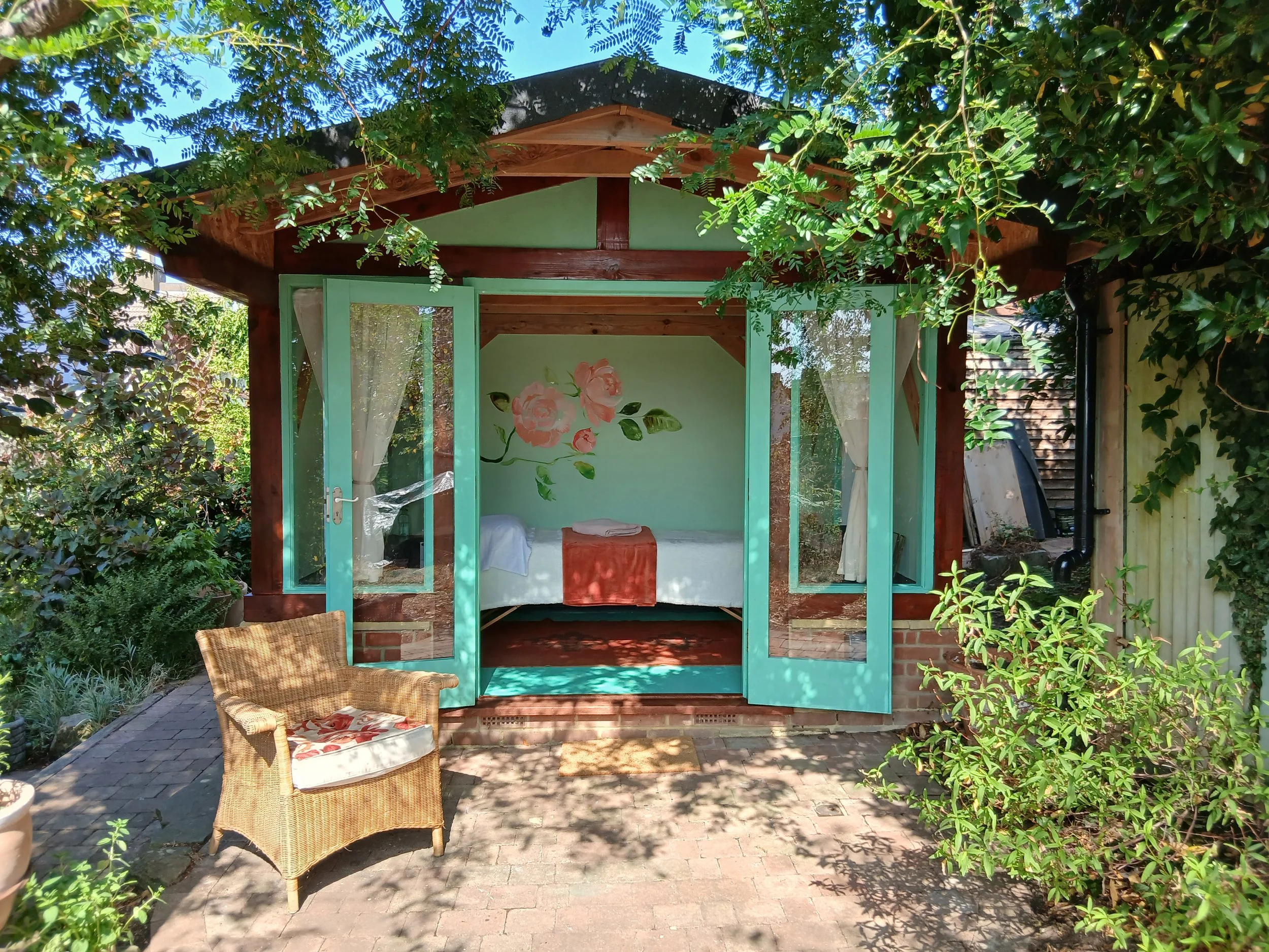 Image of the Rose Clinic from the front, a garden studio with open green French doors. There is an outdoor chair, lush greenery, and trees surrounding the area, with sunlight filtering through the leaves.