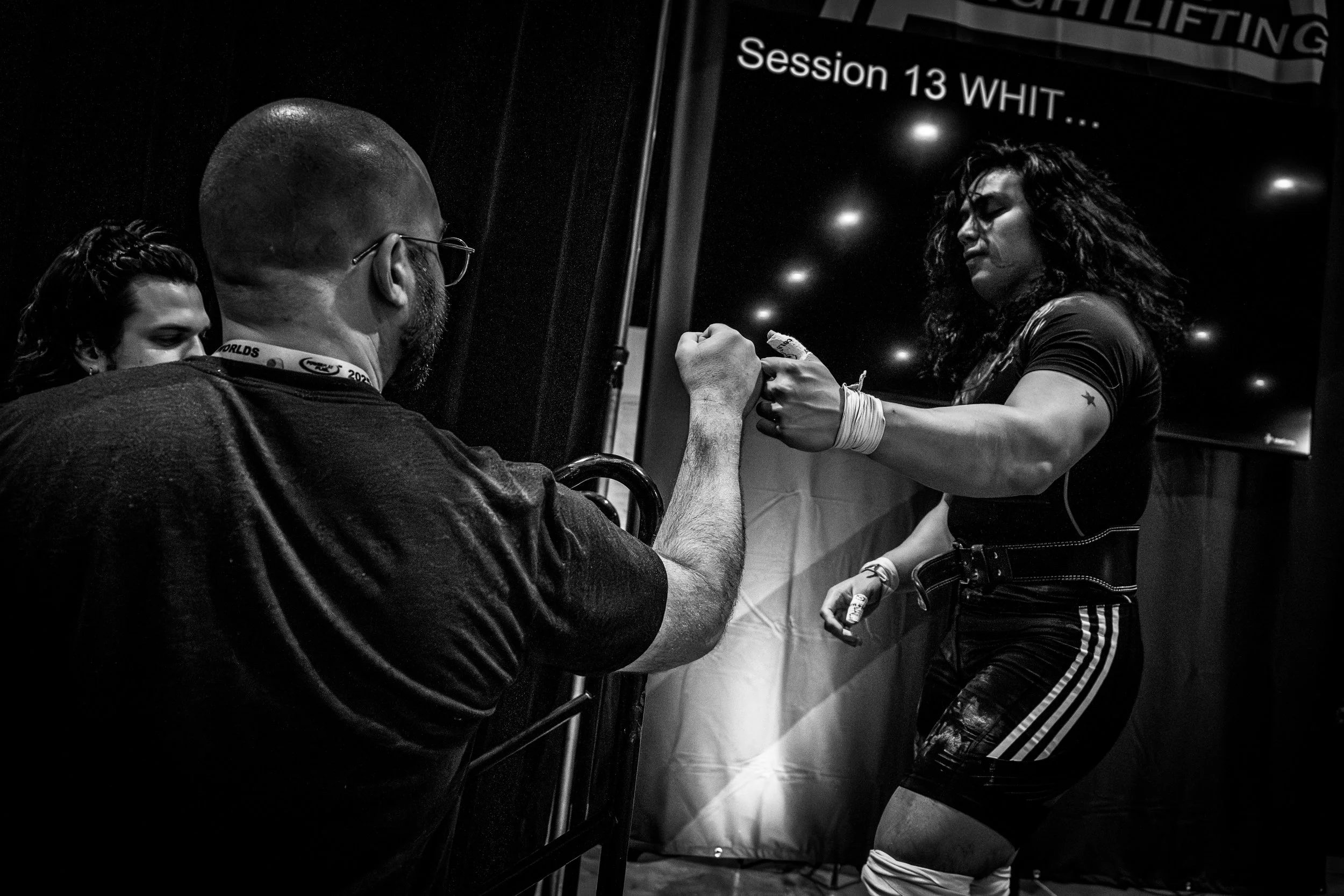 A woman athlete with curly hair and tattoos on her arm is fist bumping a man with a beard, glasses, and a lanyard at a strength competition. The woman is dressed in athletic gear and wrist wraps, standing in front of a black backdrop with a sign that says "Session 13 WHITE".