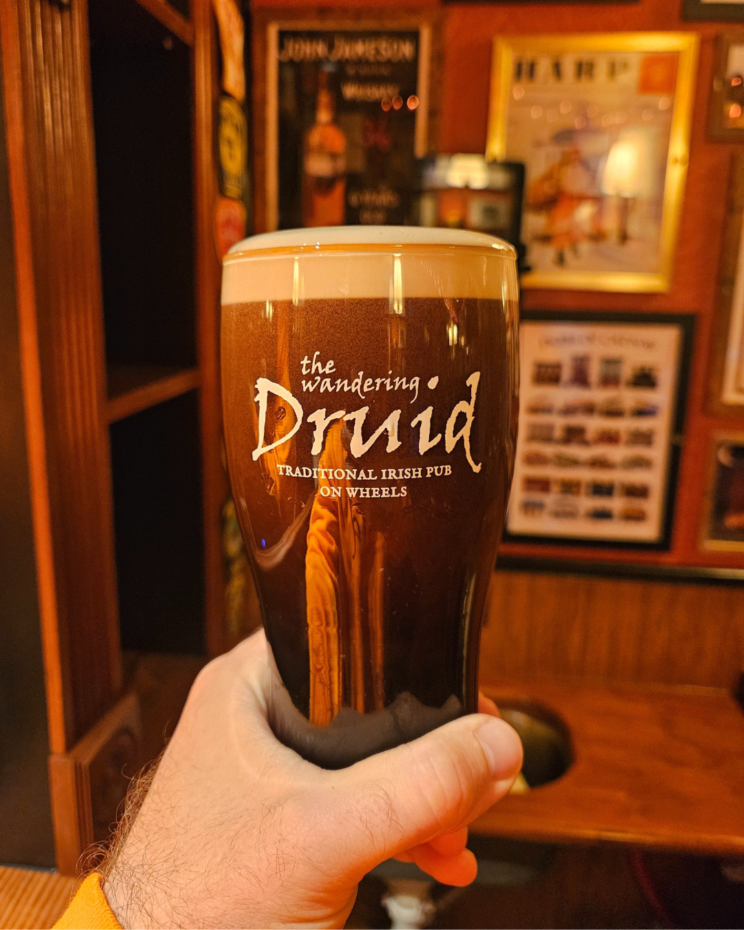 A pint glass of dark beer with a creamy head, held in a person's hand inside a Irish pub. The glass has writing that says 'the wandering Druid, Traditional Irish Pub on Wheels.' The background features wooden walls with framed pictures and posters.