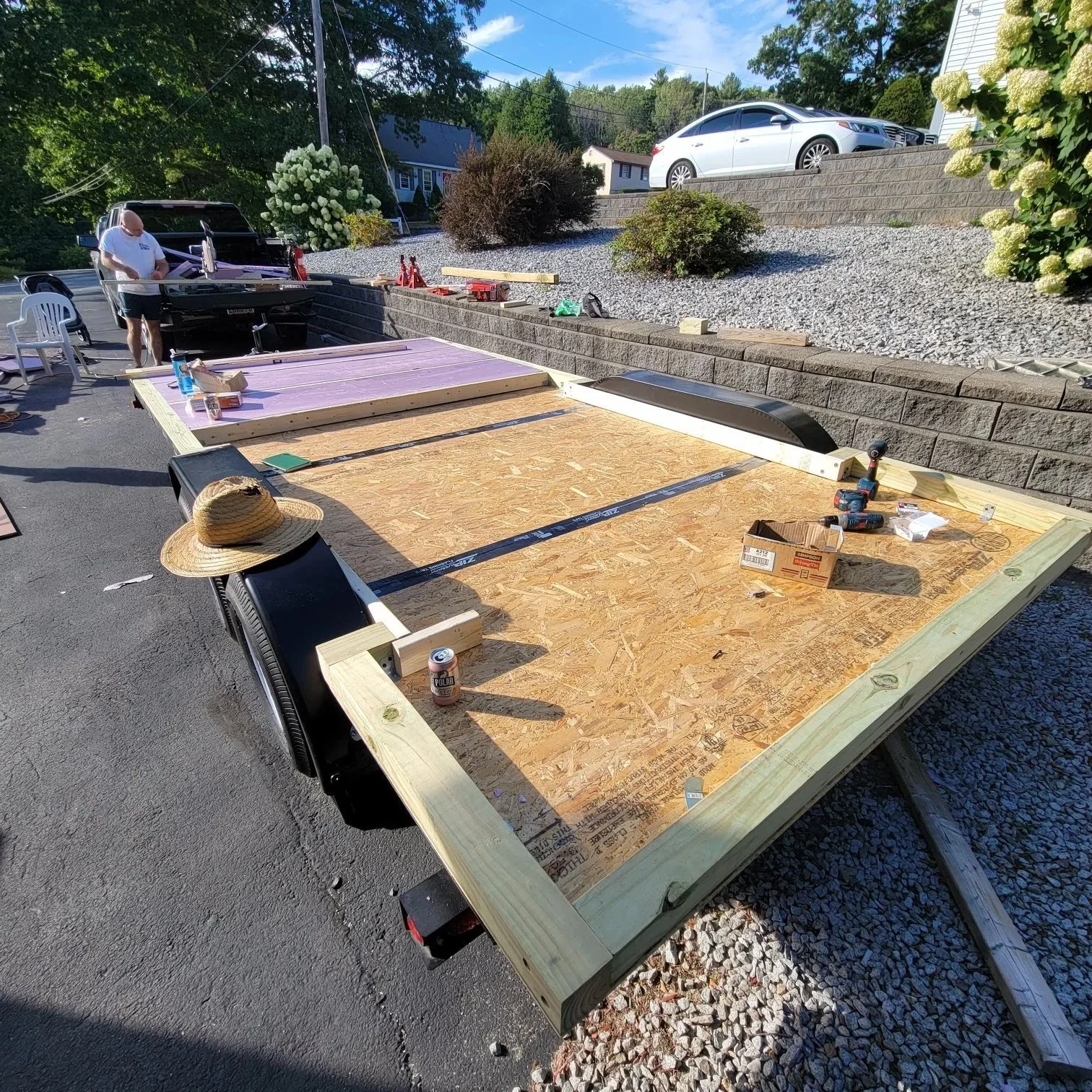 People constructing a wooden platform or deck outdoors on a sunny day, with tools and materials scattered around.