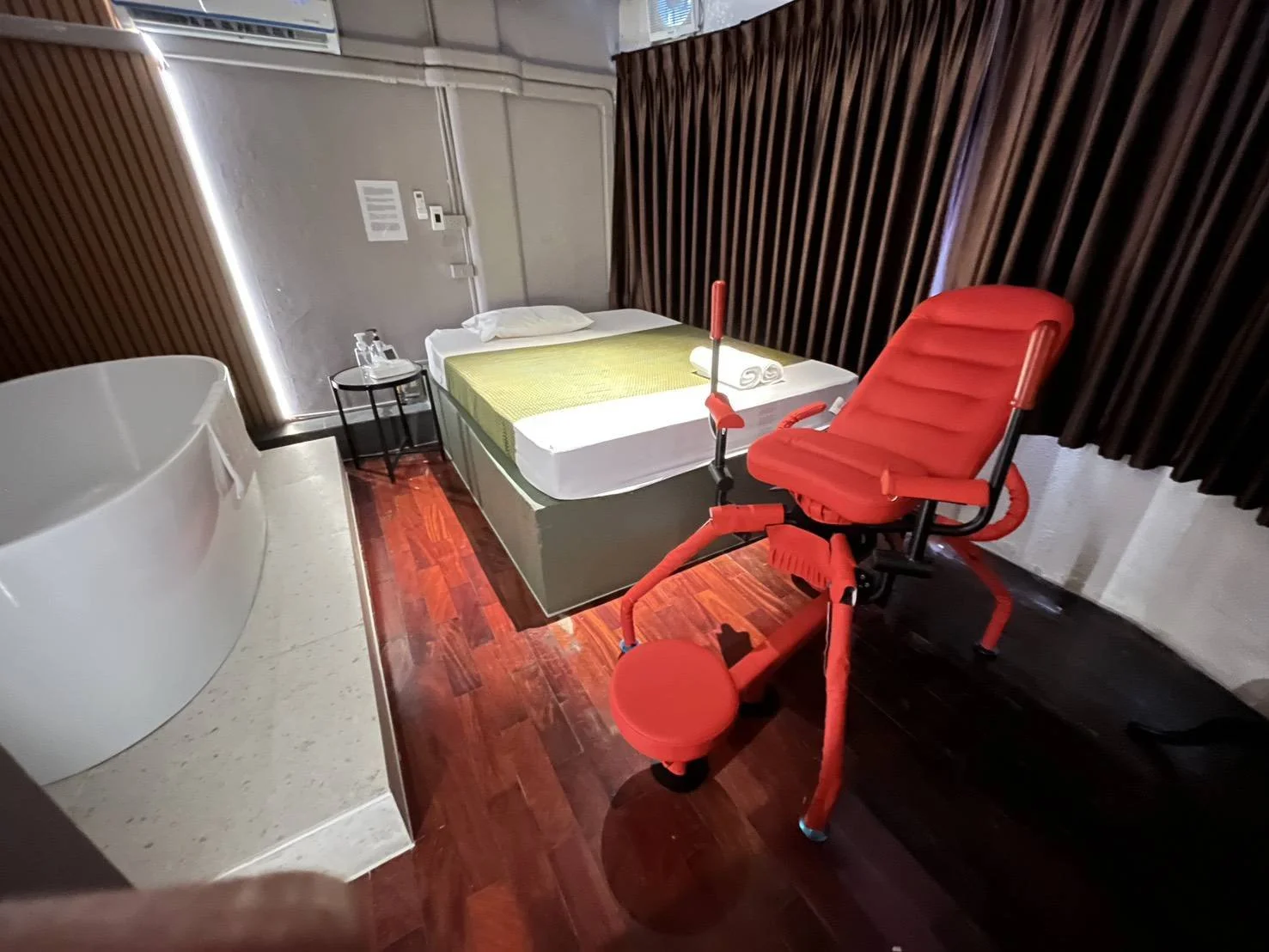 Hotel room with a white bathtub on a raised platform, a love chair in front of it, a bed with rolled towels, a window with brown curtains, and a nightstand with toiletries and a phone.
